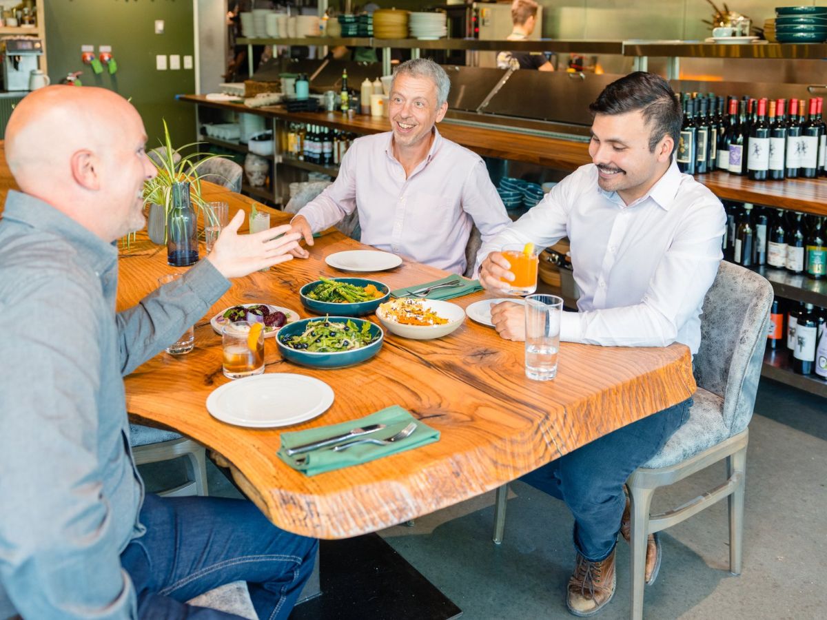 Three men sitting at a wooden table in a restaurant, talking and smiling while having lunch. The table has plates of food, drinks, and green napkins. The background shows a kitchen and wine bottles.