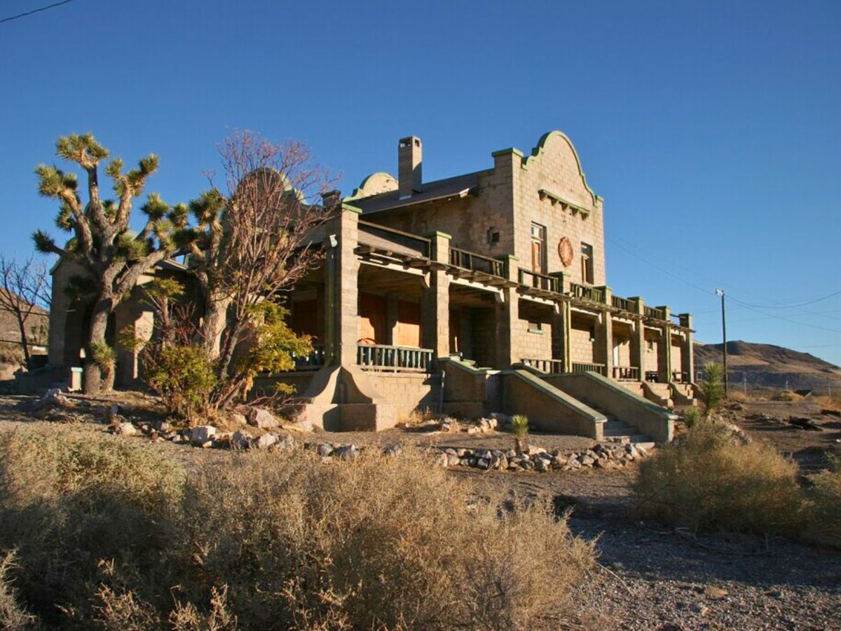 A large, rustic stone building with a porch and desert landscaping, including a prominent Joshua tree, sits under a clear blue sky.