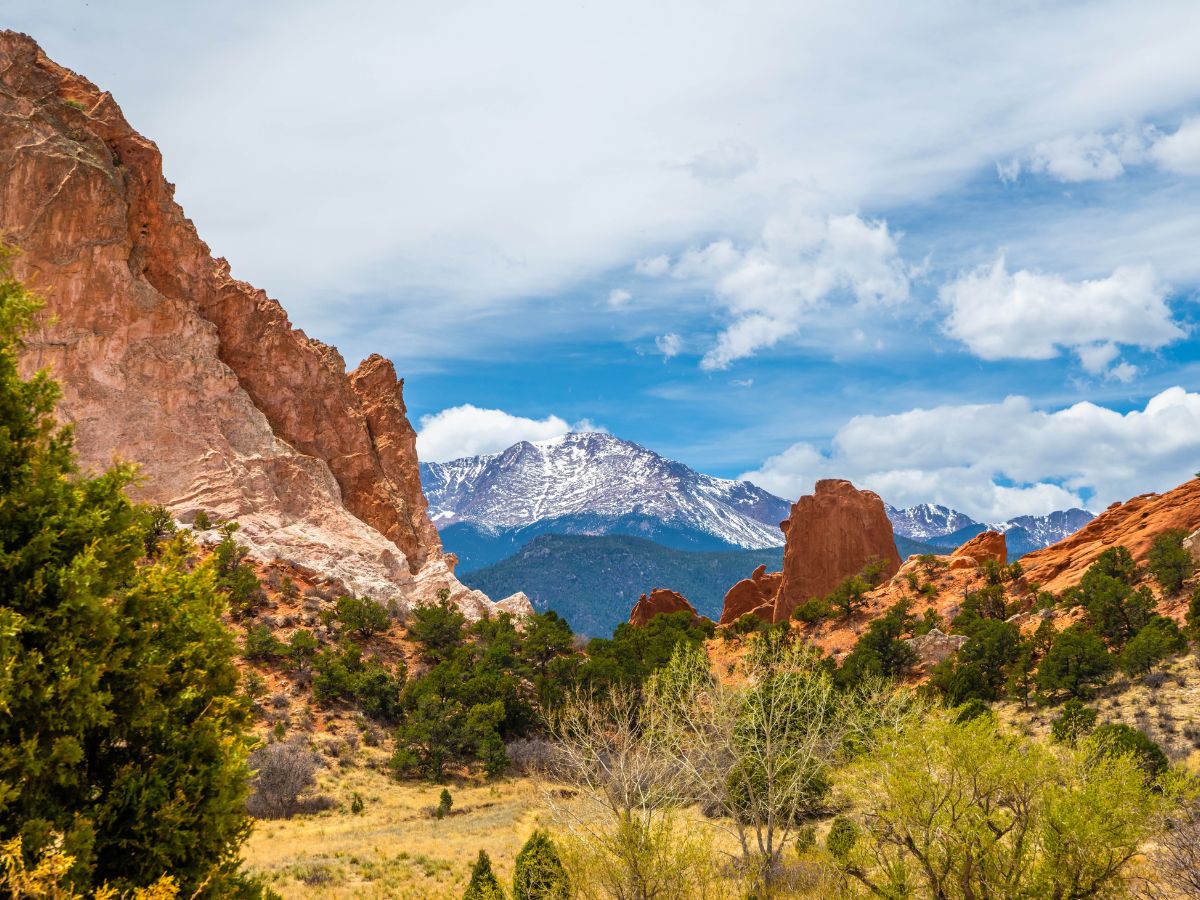 Rock formations and vegetation in the foreground with snow-capped mountains under a cloudy sky in the background.