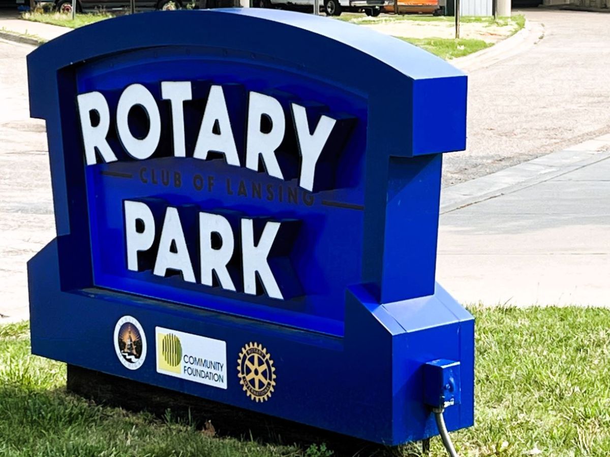 Blue sign with white text reading "Rotary Park Club of Lansing," located on a grassy area by a paved path. Logos of a local firefighting organization, a community foundation, and Rotary International are on the base.