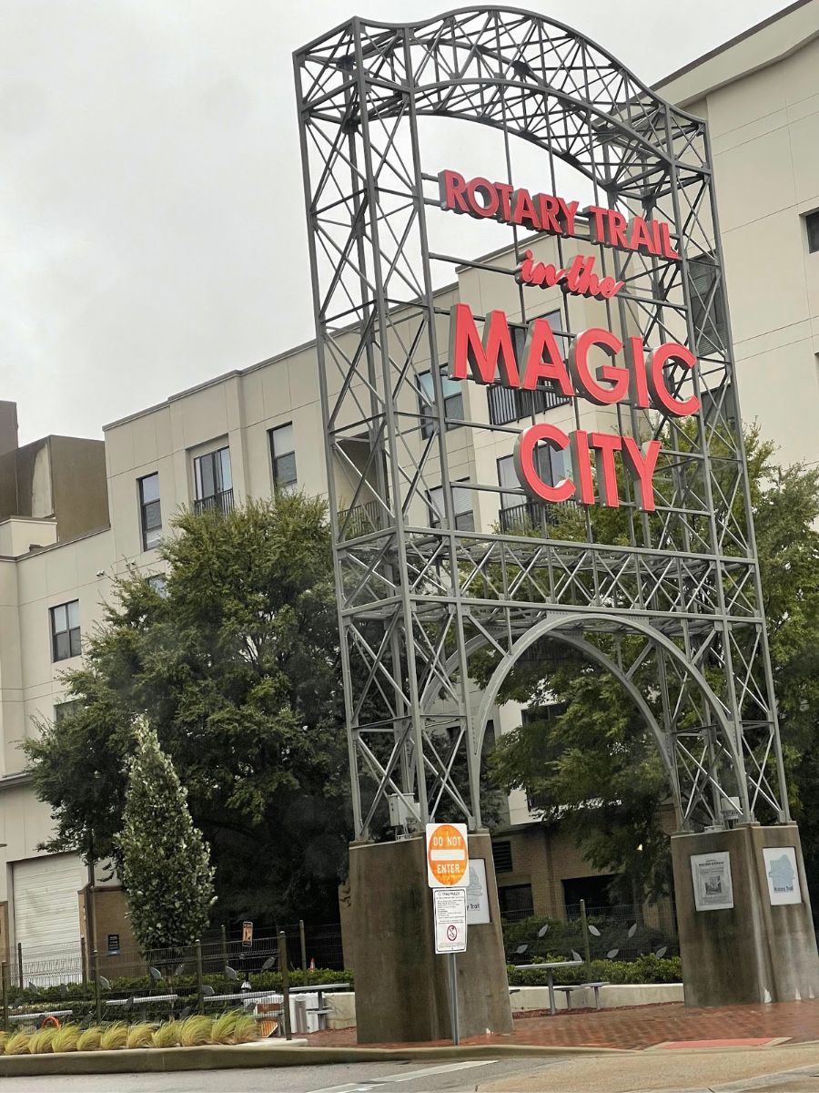 Photo of a sign reading "Rotary Trail Magic City" in front of buildings and trees. The sign is made of metal with bold letters in red and white.