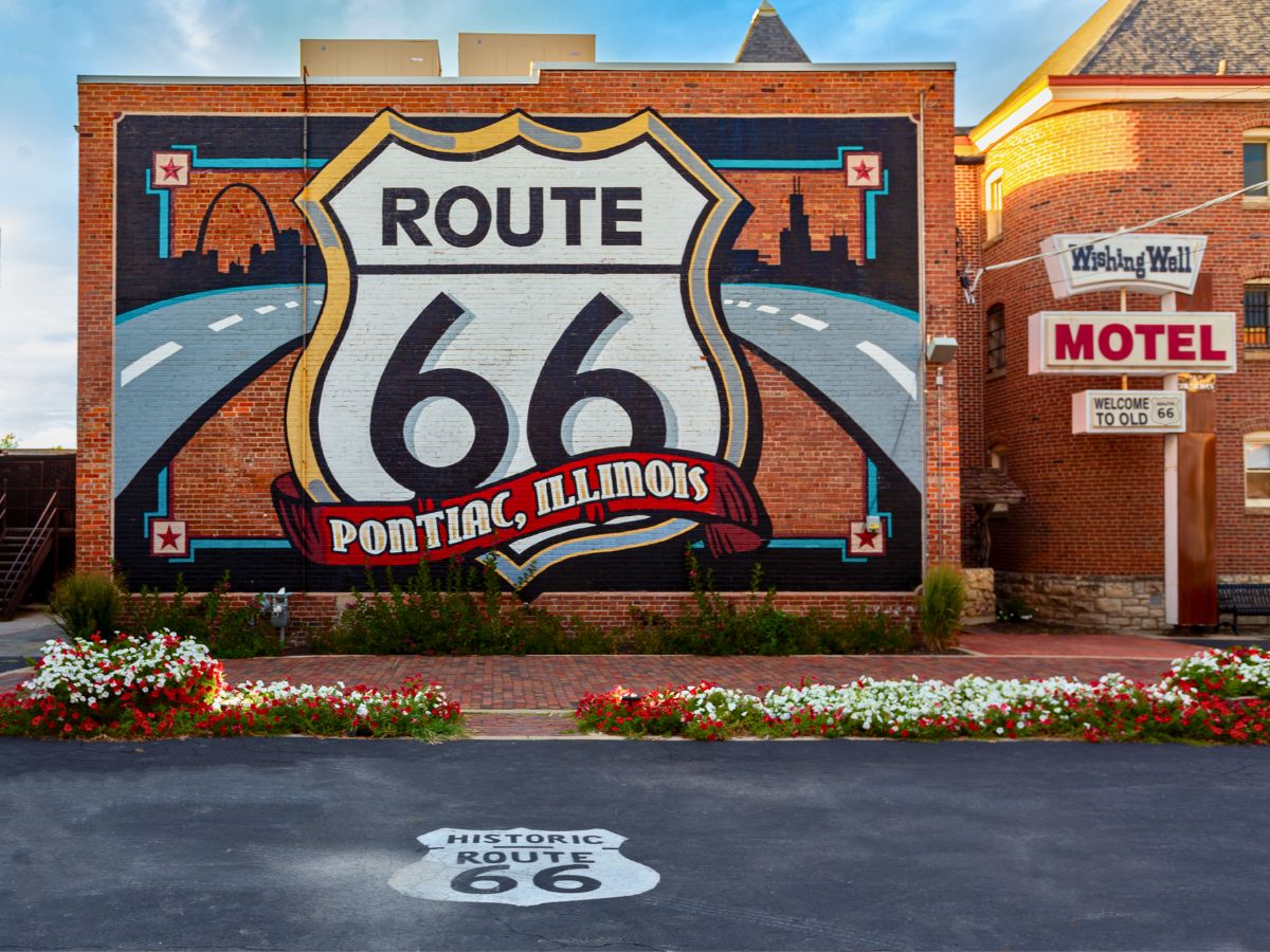 Mural of Route 66 shield with "Pontiac, Illinois" on a brick building. Nearby, a sign reads "Wishing Well Motel" with flowering plants below and a marked road in front.