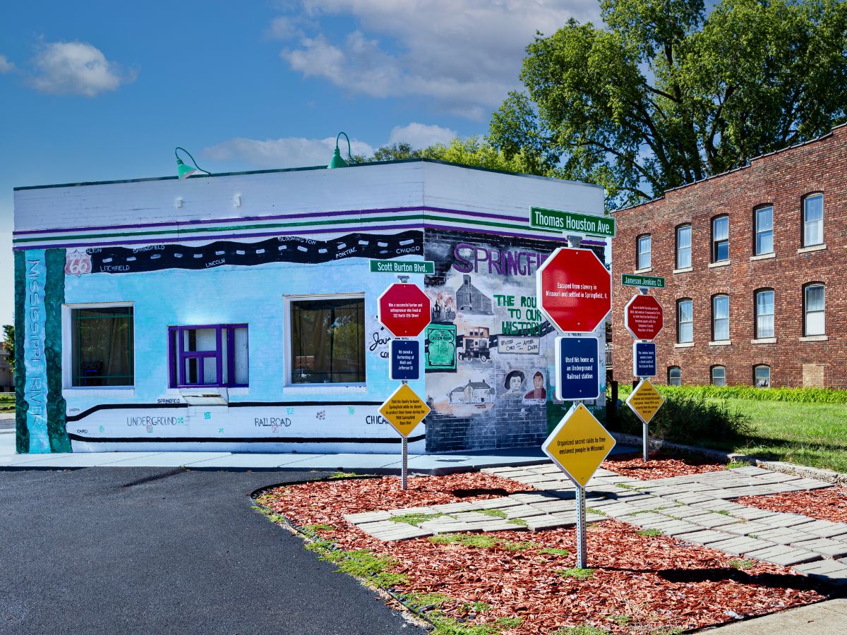 Street corner with a mural on a building and multiple colorful signs on the sidewalk. Green street signs indicate "Thomas Hudson Ave" and "Scott Dorte Blvd.