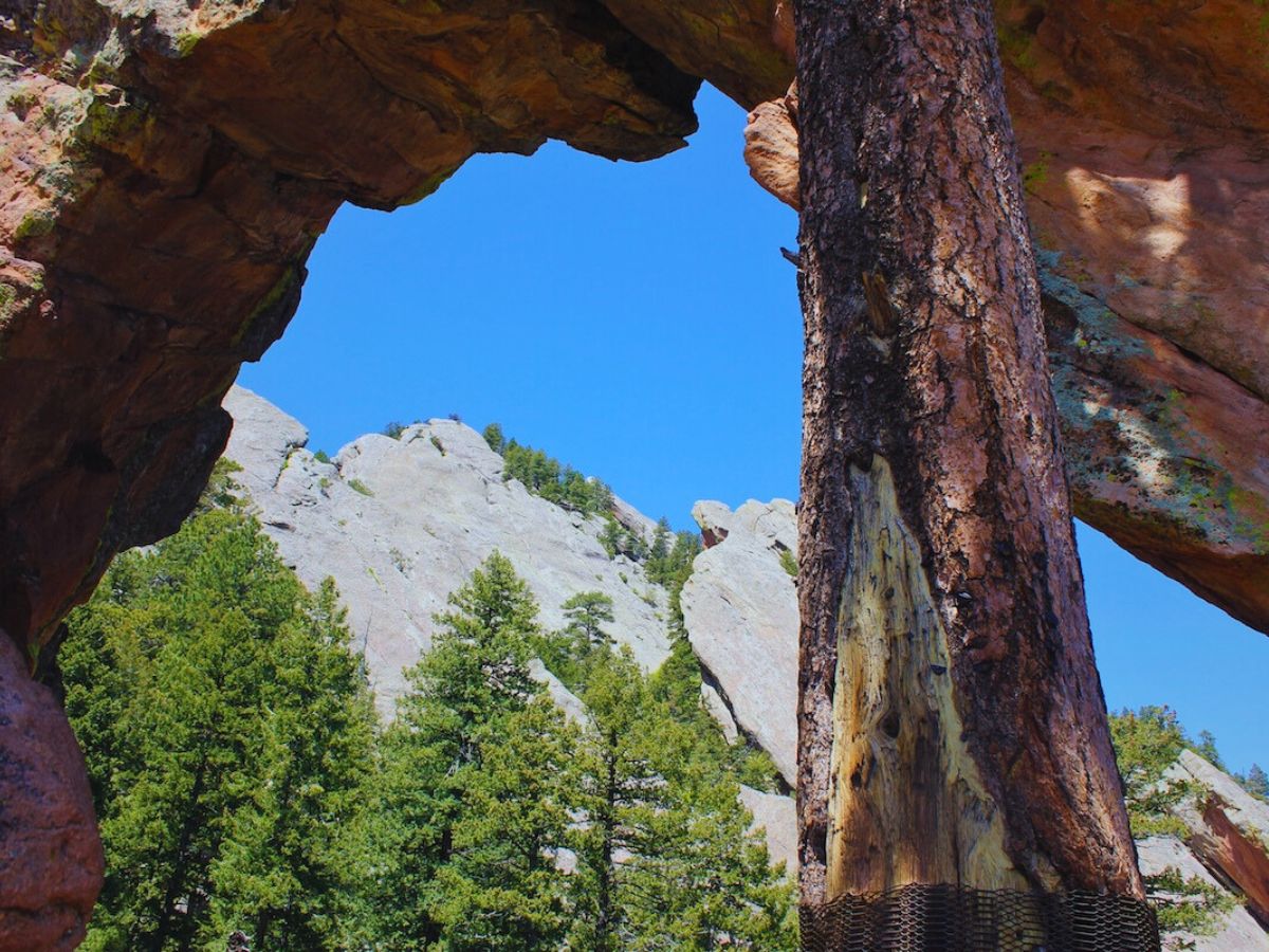 A natural rock arch frames a view of rugged mountains and evergreen trees under a clear blue sky.