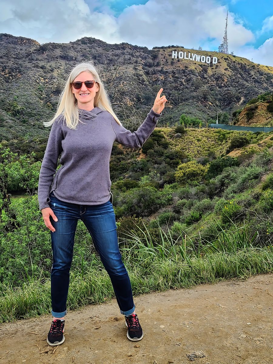 A person stands pointing at the Hollywood sign on a hill, surrounded by greenery and clouds.