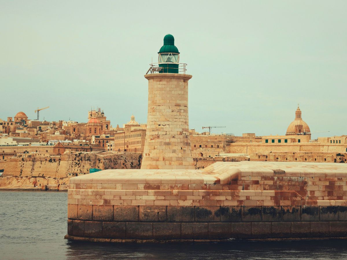 A stone lighthouse with a green top stands by the water, with an old city featuring historic buildings and a dome in the background.