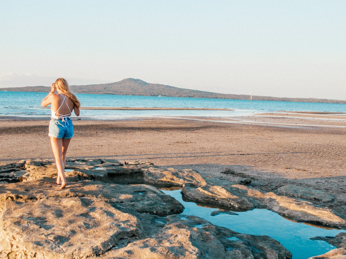 A woman in a white top and denim shorts stands on rocky terrain by the shoreline, facing the ocean, with a mountain visible in the distance.