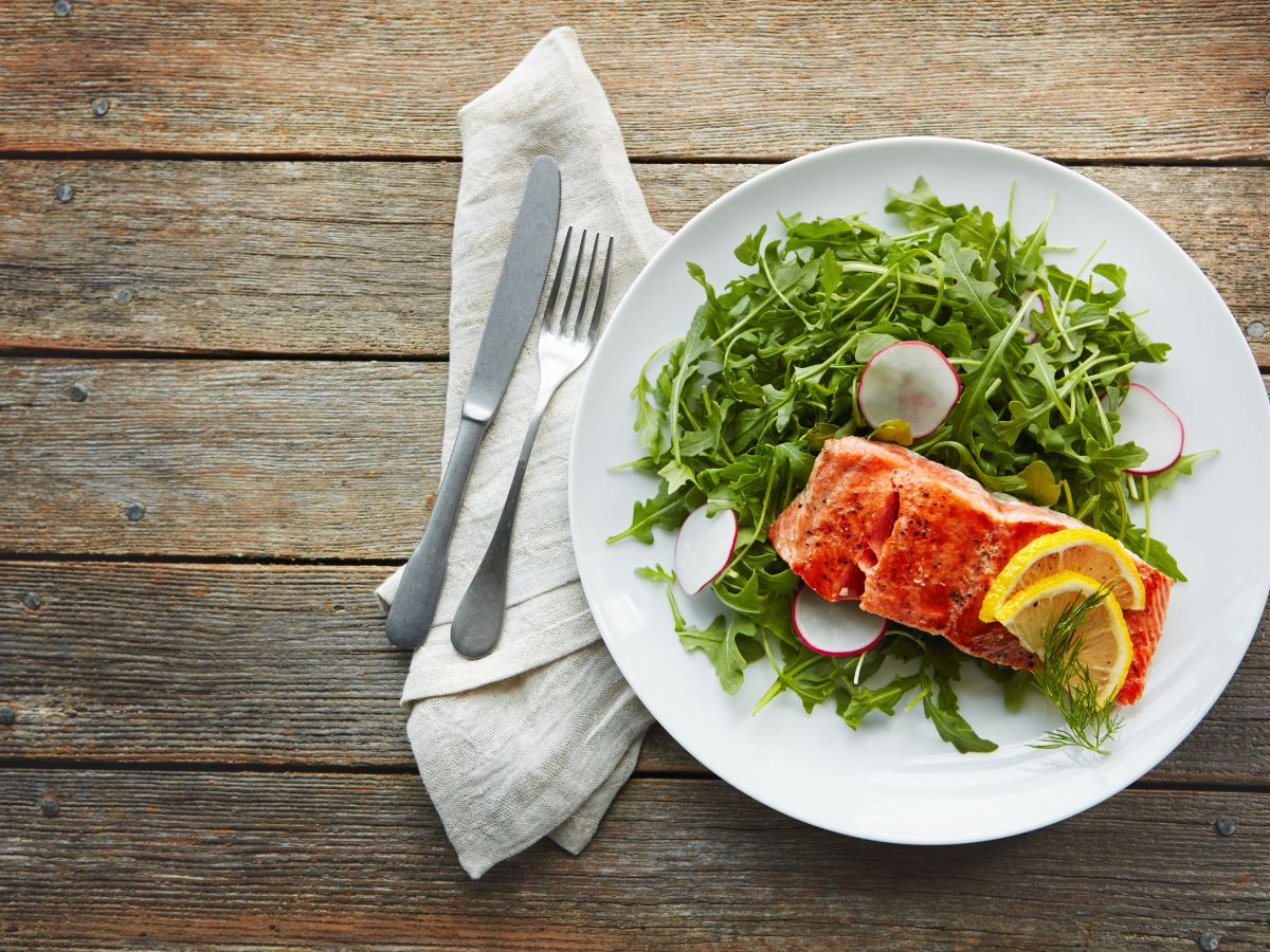 Grilled salmon fillet with lemon slices on a plate of arugula and radishes, accompanied by a fork and knife on a napkin.