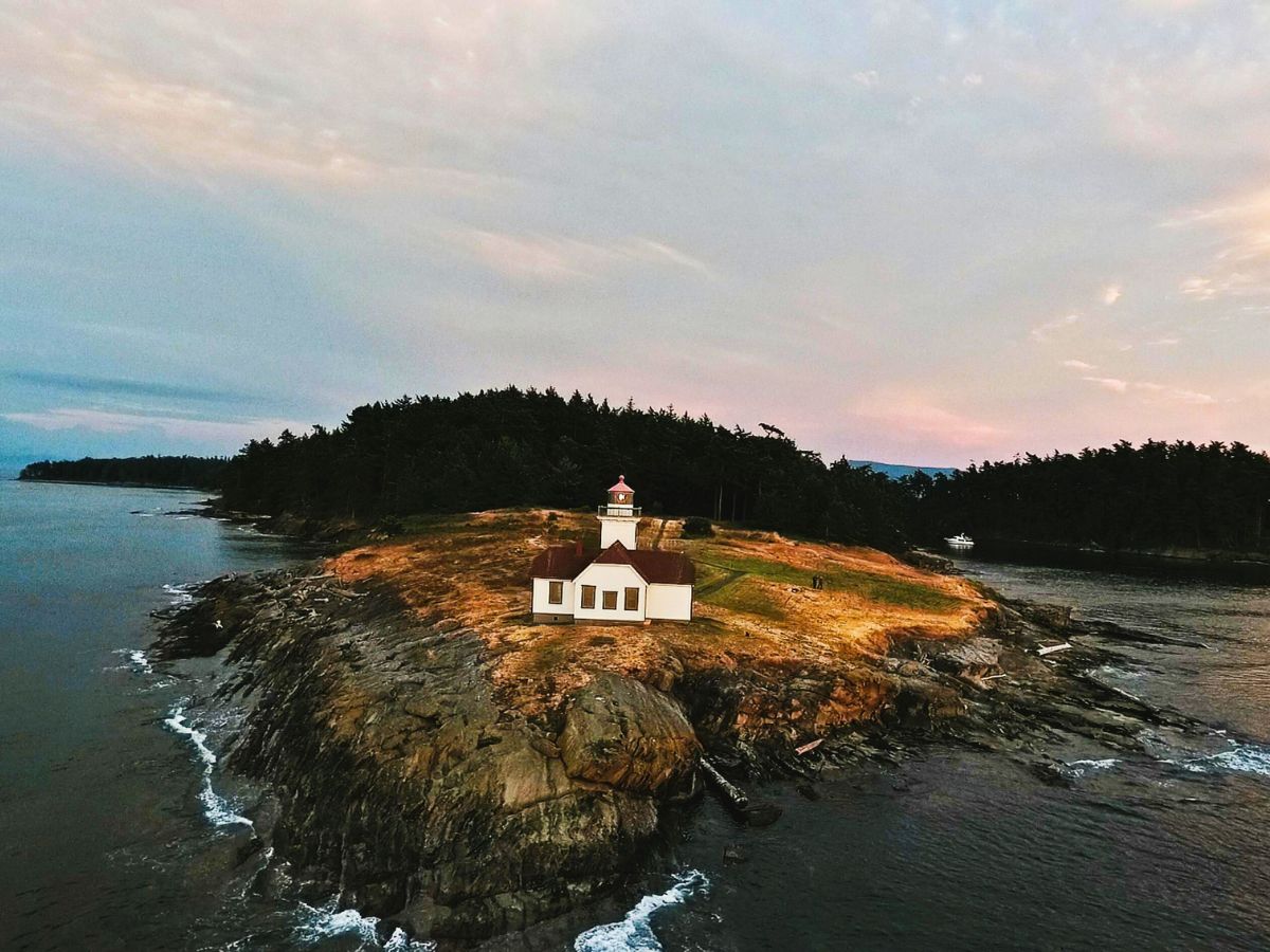 A white lighthouse stands on a rocky peninsula surrounded by water, with trees in the background under a partially cloudy sky.