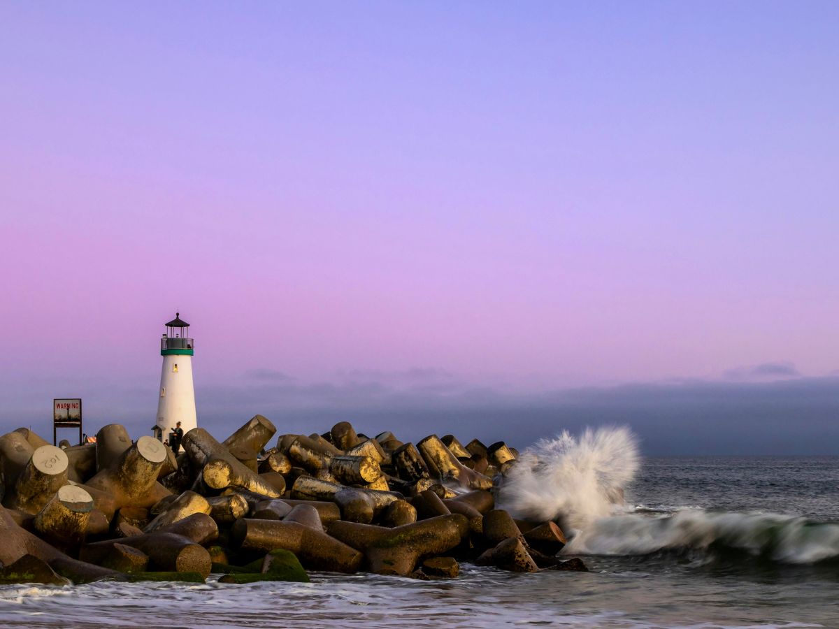 A white lighthouse stands on a rocky breakwater with waves crashing against it, set against a purple-hued sky.