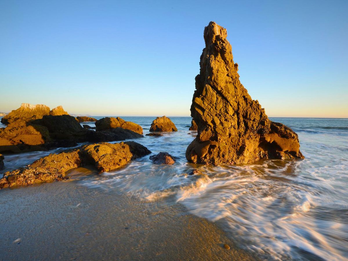 Large rocky formations stand in shallow ocean waters, with waves gently washing around them under a clear, blue sky at sunset.