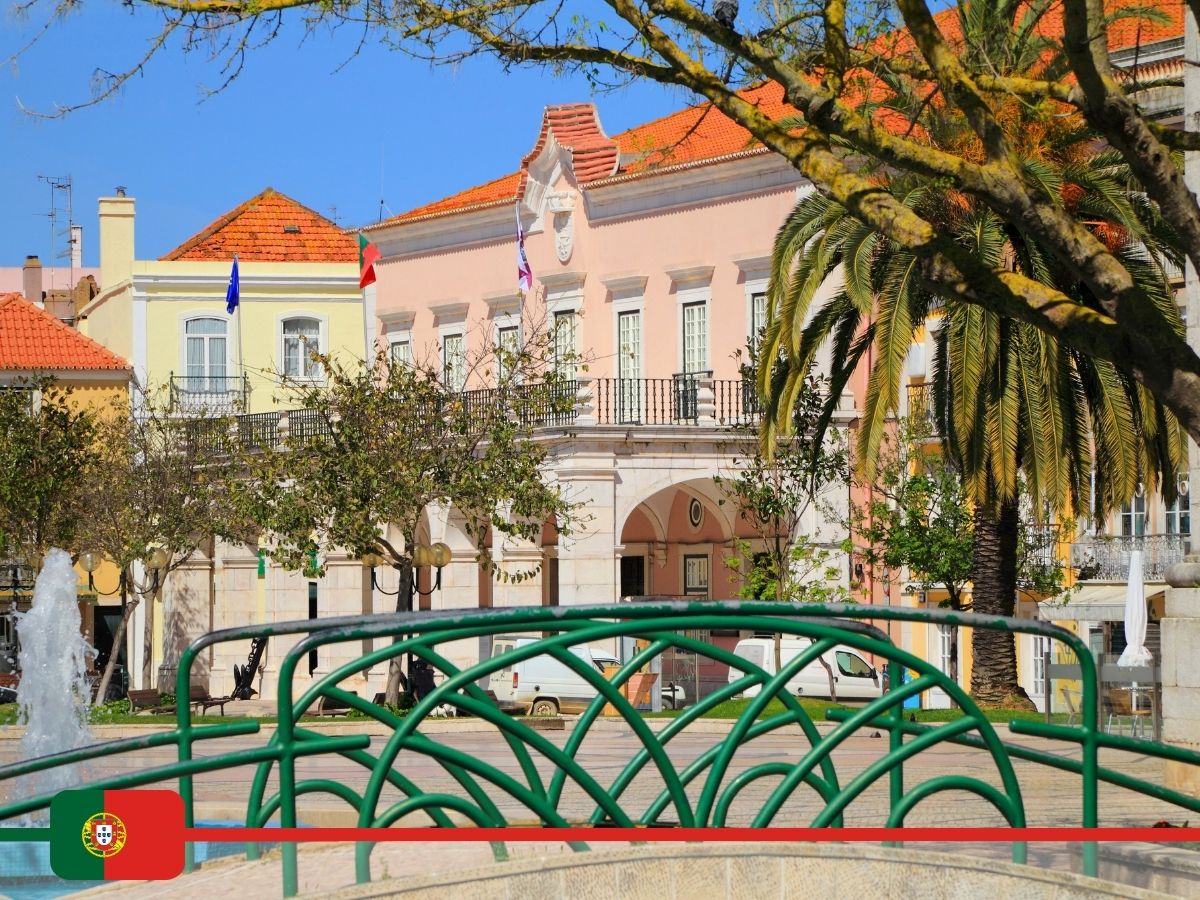 A picturesque square in Setubal, Portugal, featuring a fountain, trees, and a colonial-style building with flags and a palm tree.