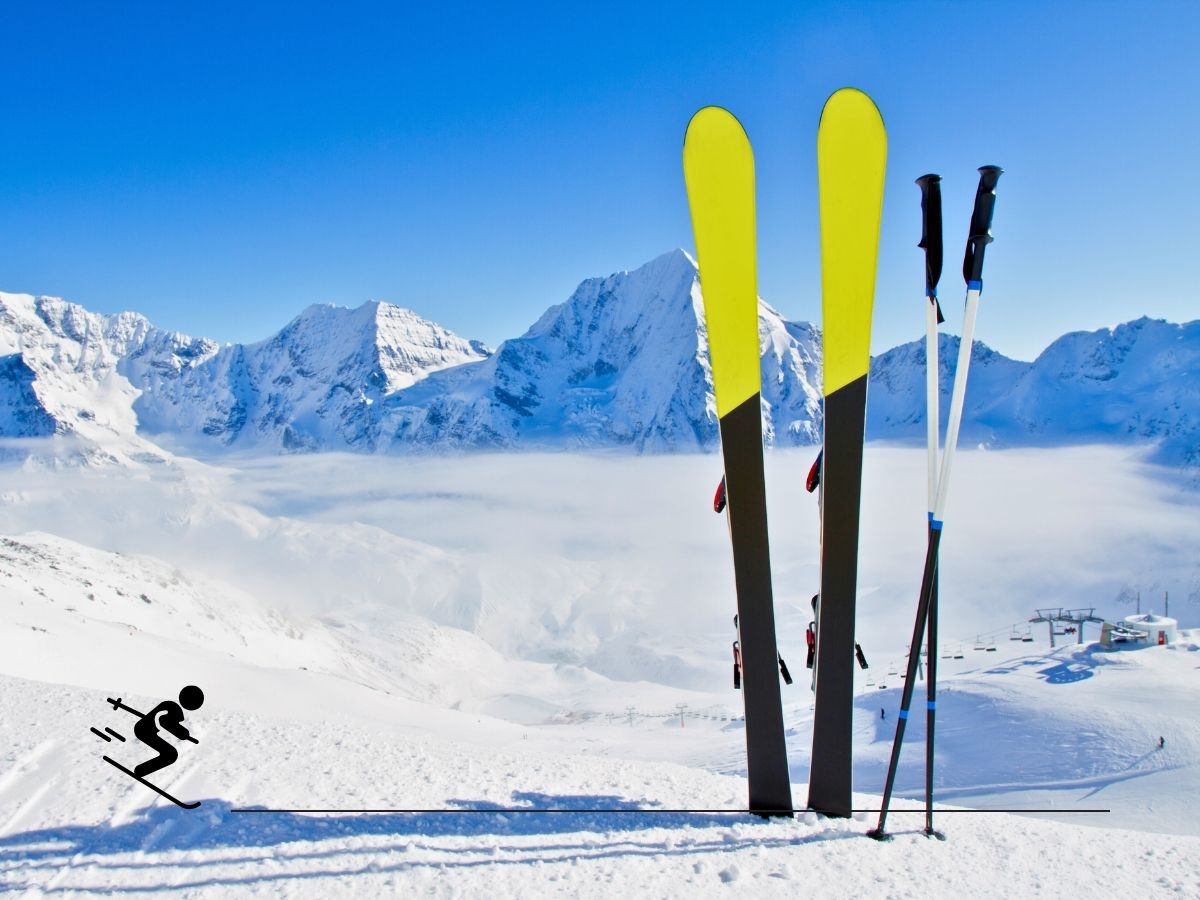 Two pairs of skis and poles standing upright on a snowy mountain slope, with misty mountains in the background under a clear blue sky.
