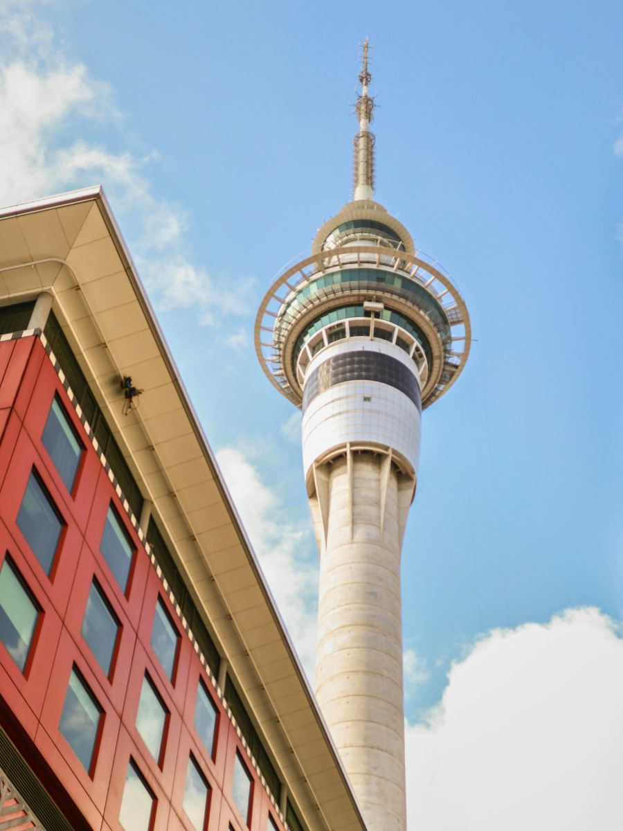 A tall concrete and glass communications tower rises into a partly cloudy blue sky, adjacent to a red and beige building with multiple windows.