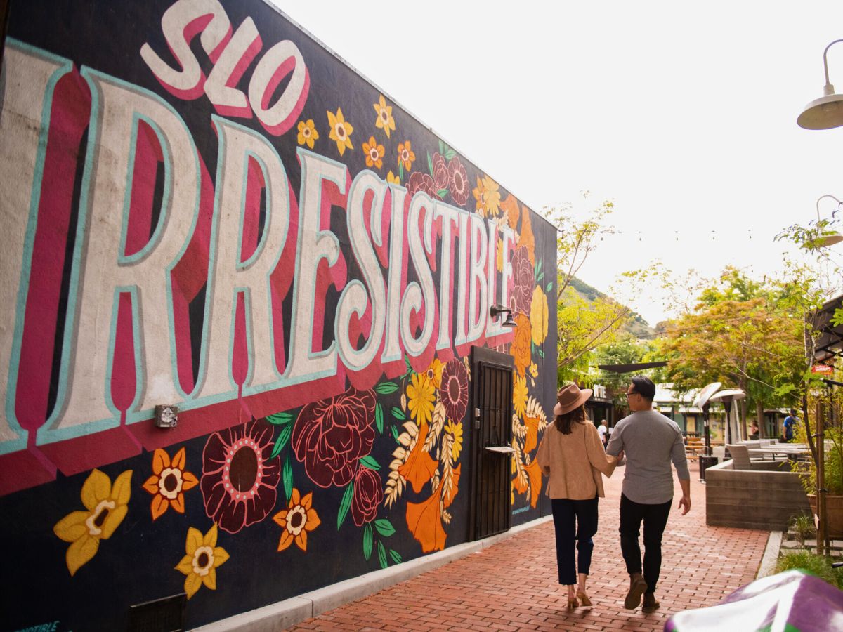 A couple walks hand in hand along a brick path beside a colorful mural with the word "IRRESISTIBLE" in large letters.
