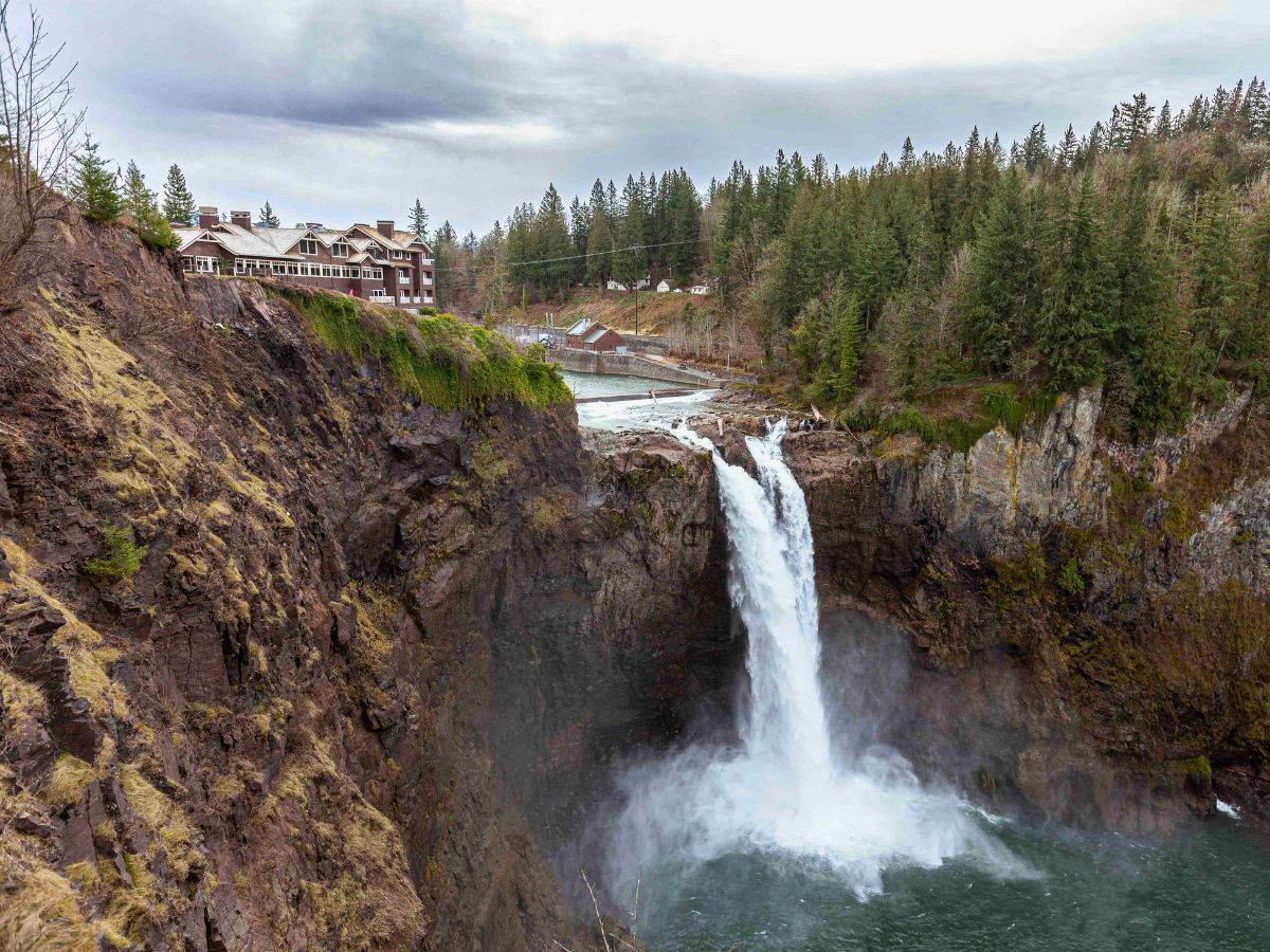 A large waterfall cascades down a rocky cliff surrounded by trees, with a building visible on the left.