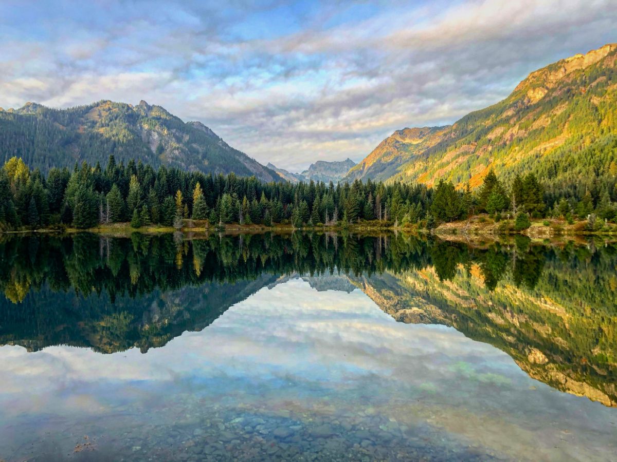 A serene mountain lake reflects the surrounding forest and rugged peaks under a partly cloudy sky.