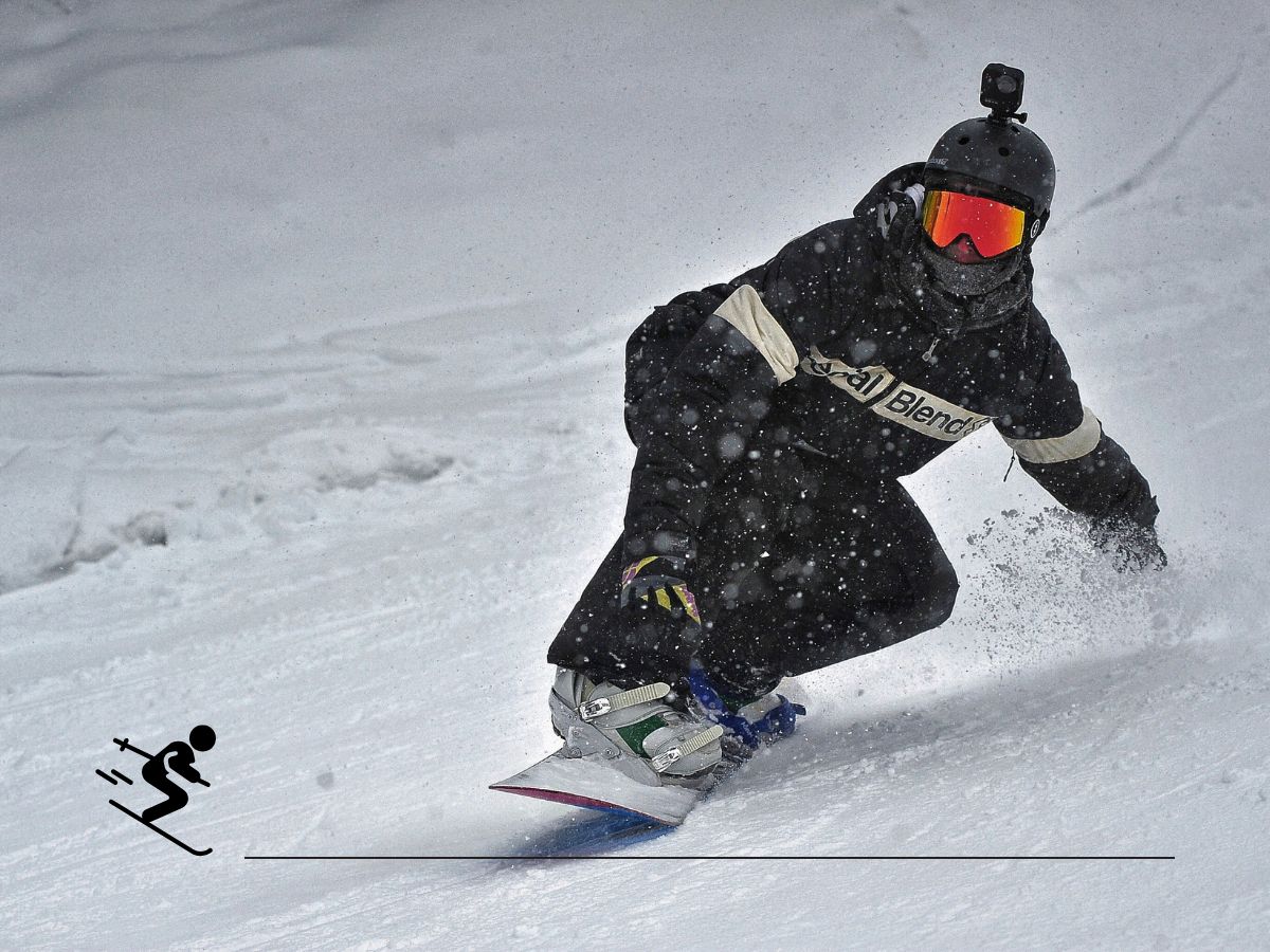 Snowboarder carving down a snowy slope, wearing dark protective gear and goggles, with a camera mounted on their helmet.