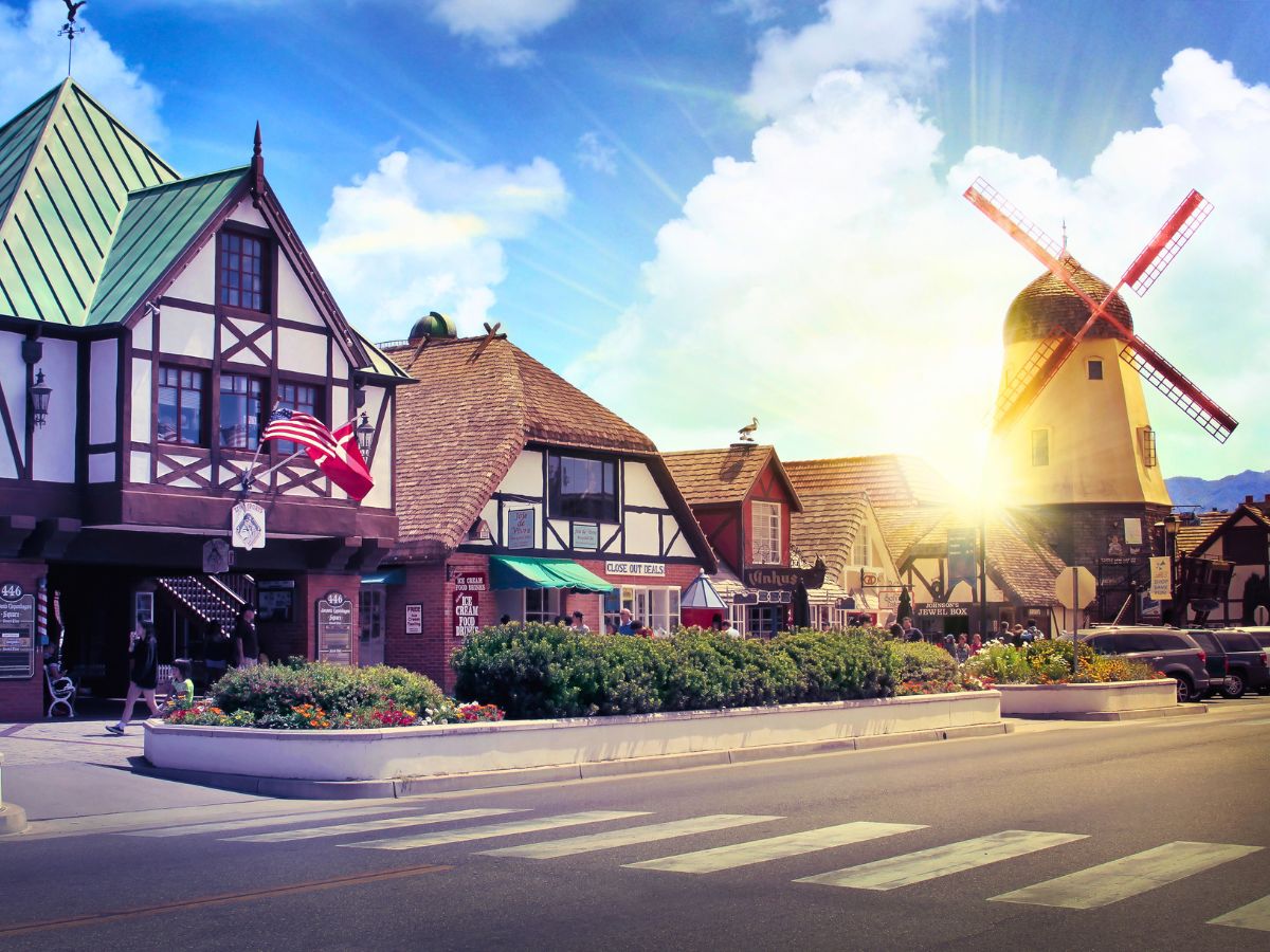 A street scene with half-timbered buildings, a windmill, and colorful plants under a sunny sky. American flags hang from one building, and several cars are parked along the road.