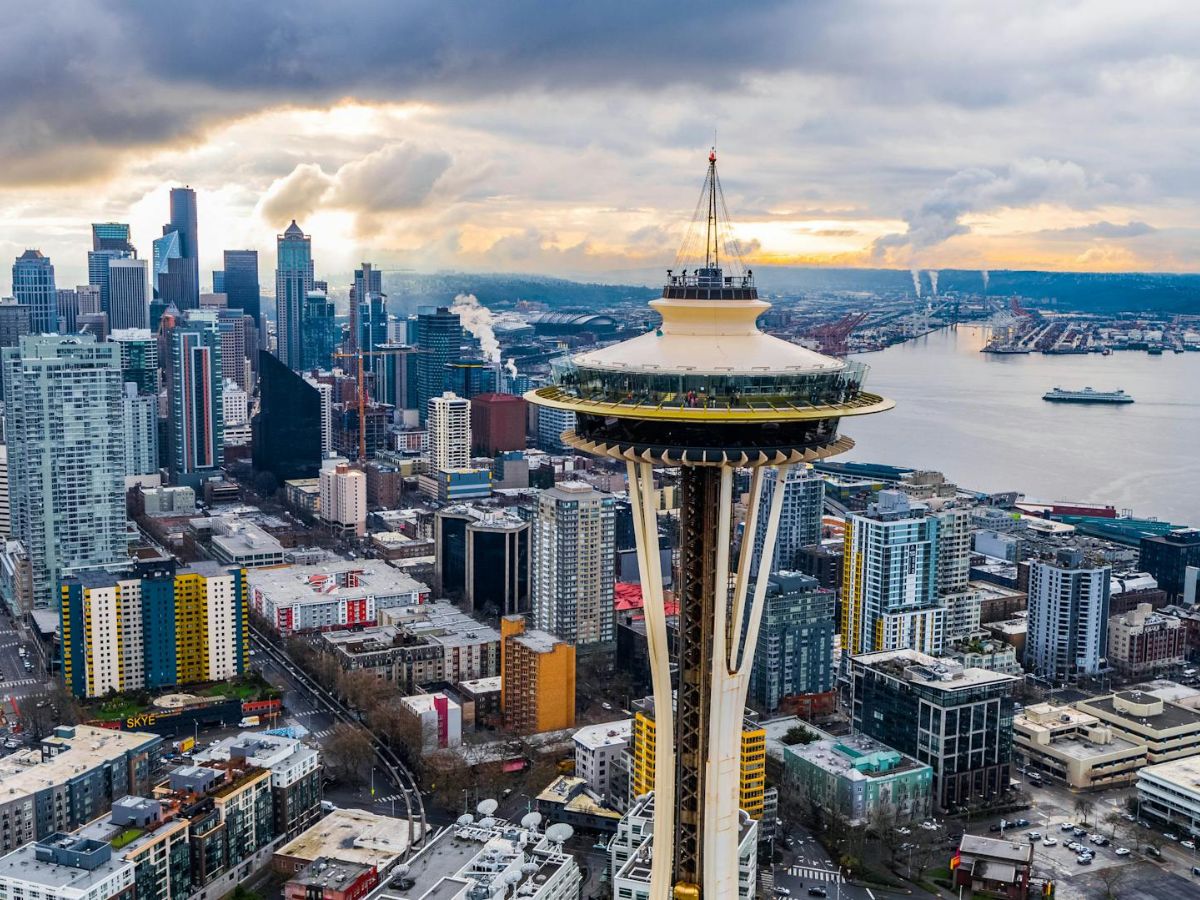 Aerial view of Seattle featuring the Space Needle in the foreground, with the city's skyline and a body of water in the background under a cloudy sky.