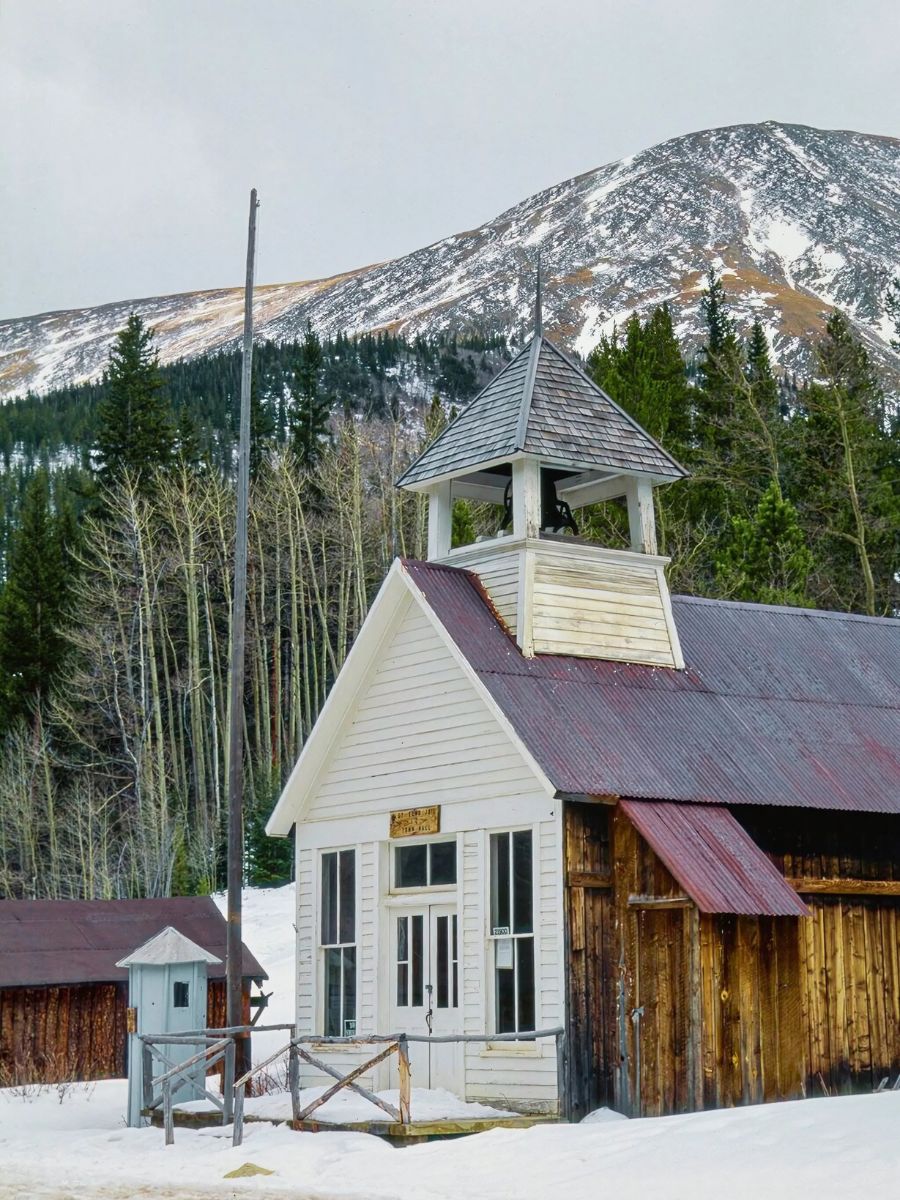 A rustic wooden church with a bell tower stands in a snowy landscape, surrounded by trees, and backed by a mountain.