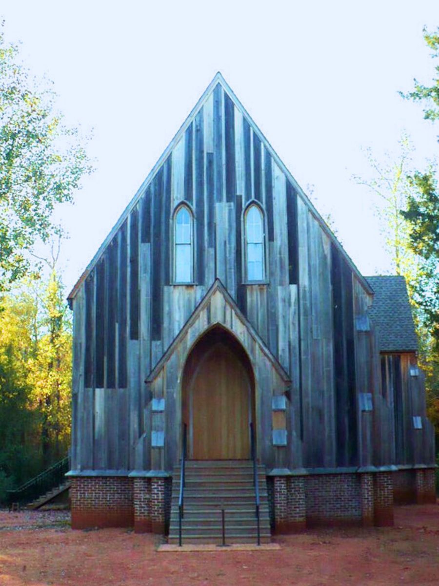A quaint, old wooden church with a steep triangular roof and two tall windows, set among trees. The front entrance has a small set of stairs.