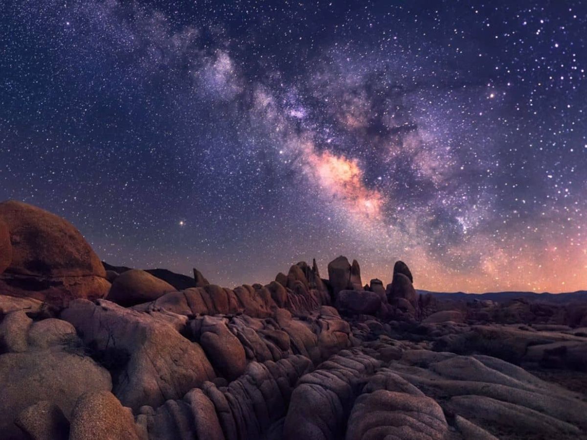 A night sky over a rocky desert landscape, featuring a clear view of the Milky Way galaxy with numerous stars scattered across the sky.