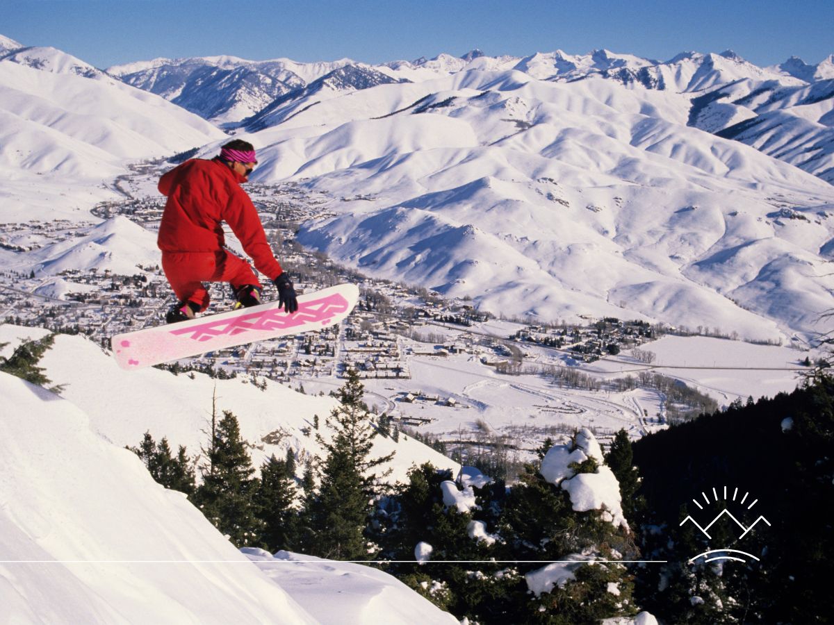 Snowboarder in red gear mid-air on a snowy mountain with a snowy valley and town in the background. Bright and sunny weather.