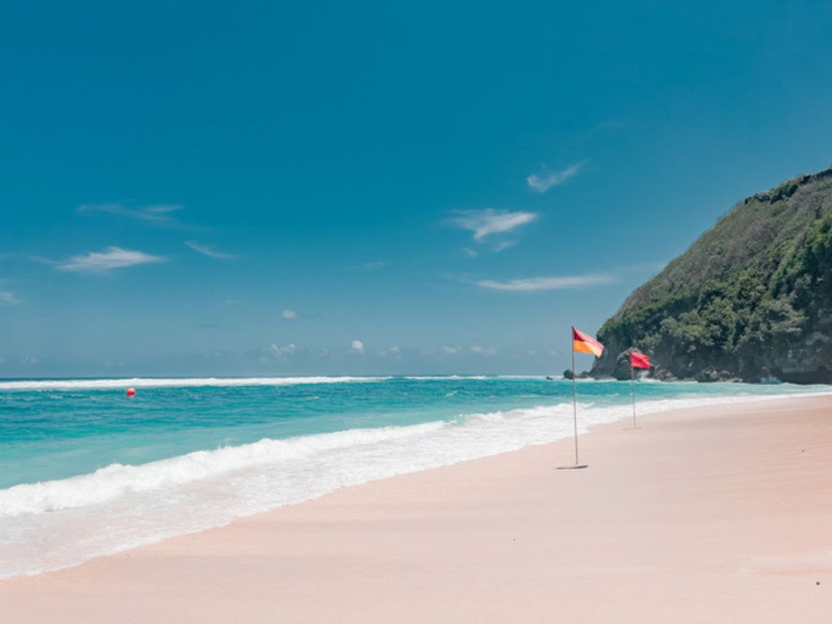 A beach scene with clear blue water and sky, featuring two safety flags in the sand. A grassy cliff is visible on the right side of the image.