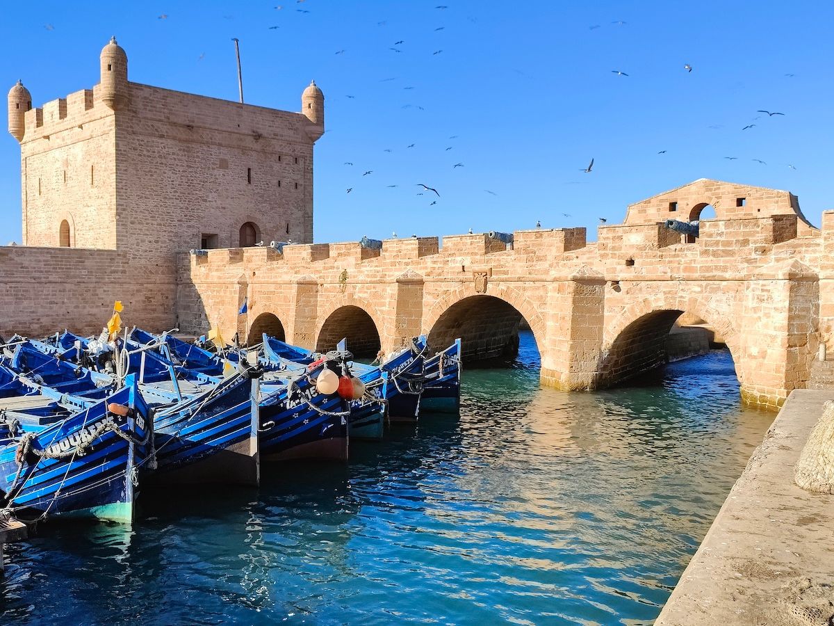 A historic stone harbor with an arched bridge, docked blue boats, and a defensive tower under a clear blue sky with seagulls flying overhead.