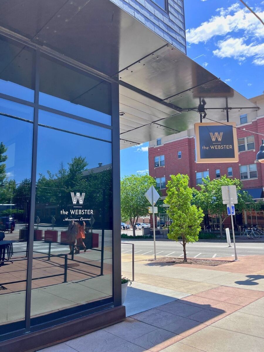 Exterior of "The Webster" storefront on a sunny day, with glass windows reflecting the street and a sign hanging above. Trees and buildings are visible in the background.