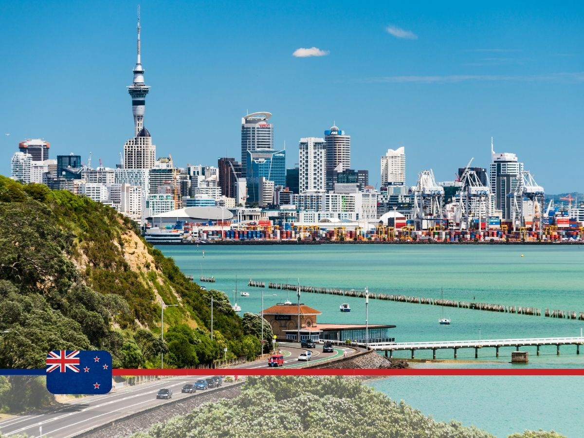 View of Auckland skyline with Sky Tower and various buildings, harbor, and waterfront activity on a clear day. The New Zealand flag and a red and blue ribbon are in the bottom foreground.