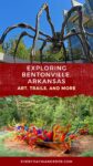 A sculpture of a spider and colorful glass artwork are featured outside a building, promoting Bentonville, Arkansas with text highlighting art, trails, and more.