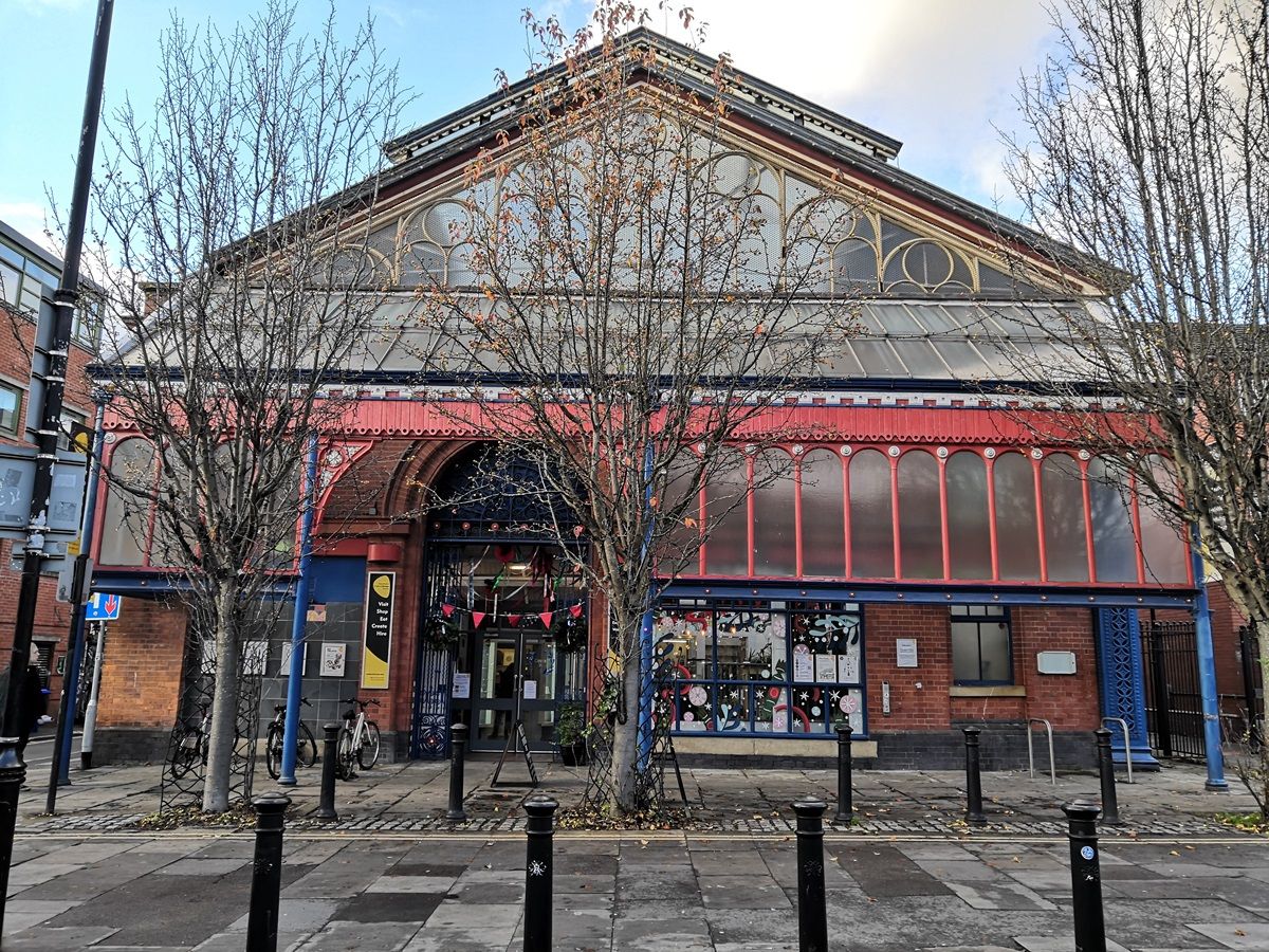 A historic building with a glass roof and a red-painted façade, featuring large front windows and a decorated entrance, stands behind leafless trees on a cobblestone sidewalk.