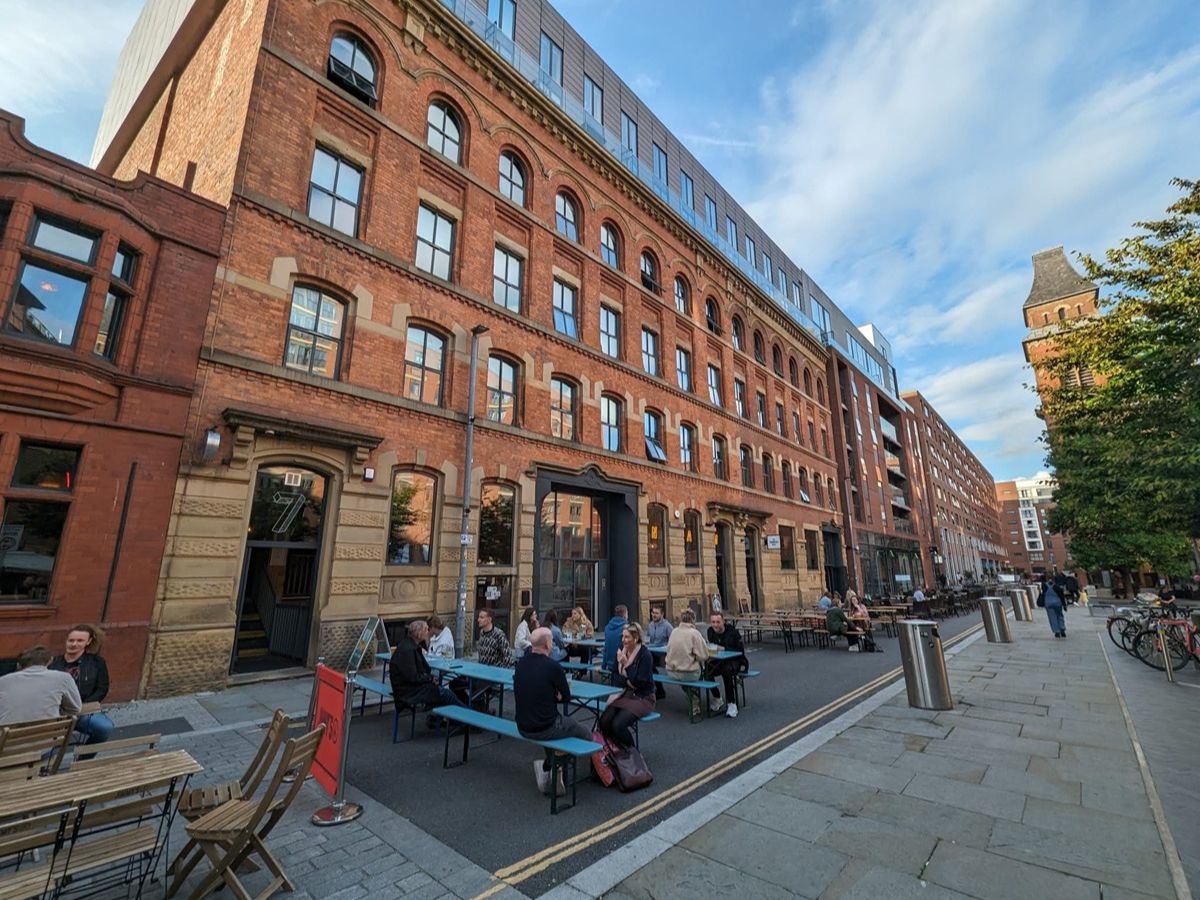 People sitting at outdoor tables along a narrow street, with a large brick building and additional structures visible in the background, during a clear day.