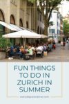 People sitting at outdoor cafe tables in a street surrounded by historic buildings and trees in Zurich.