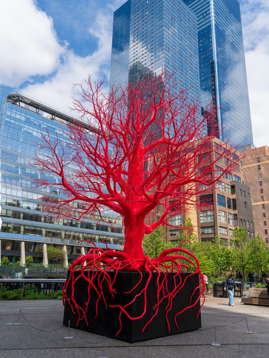 A bright red tree sculpture with visible roots stands in front of modern glass skyscrapers and buildings under a partly cloudy sky.