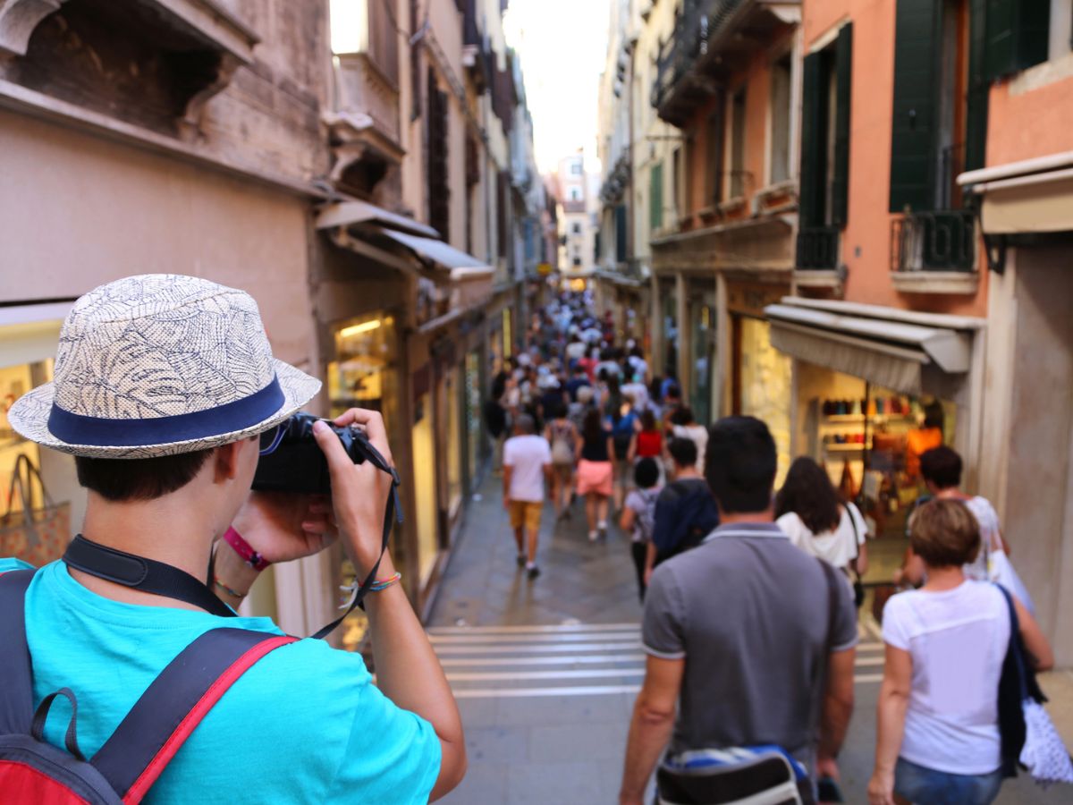 A person in a hat takes a photo of a crowded, narrow street lined with shops and buildings, with blurred people walking ahead.