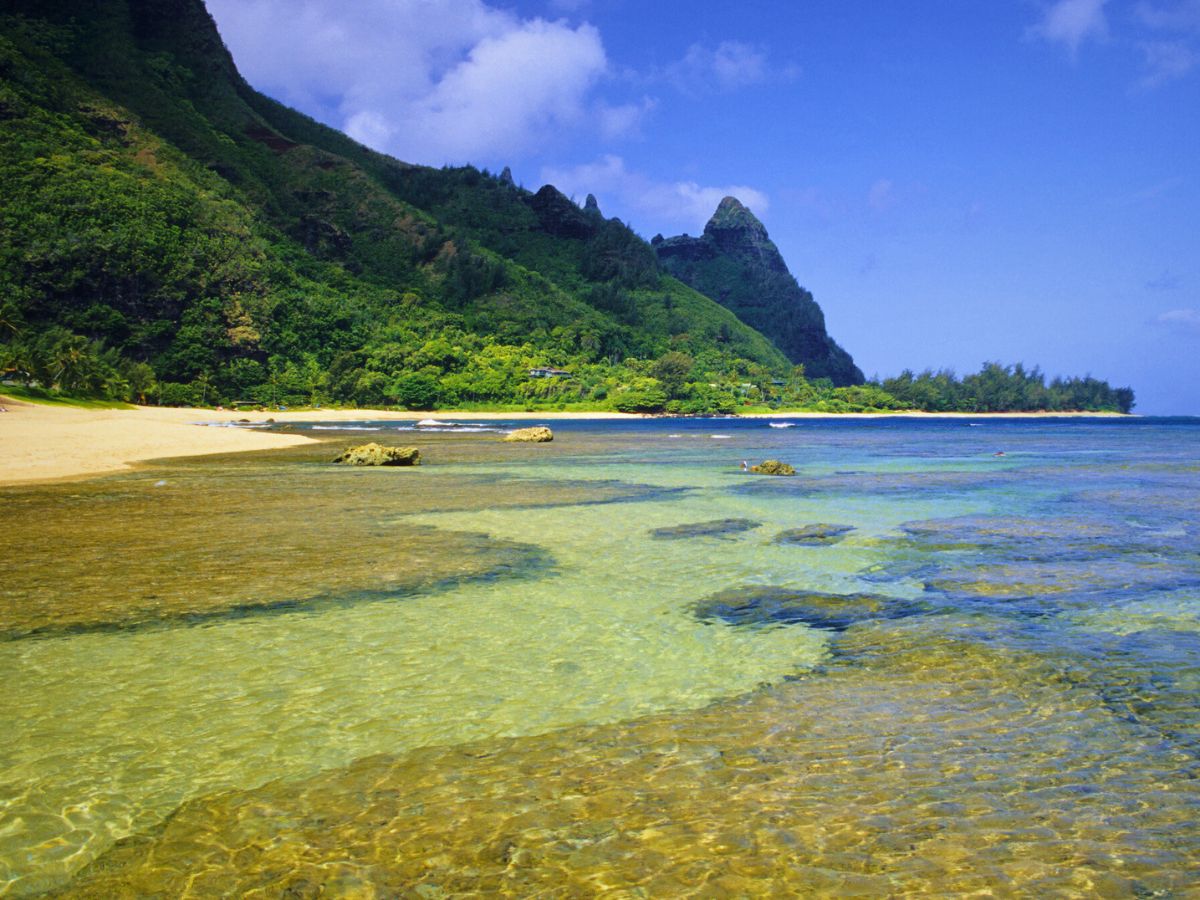 A tropical beach with clear turquoise water, rocky outcrops, and lush green mountains under a blue sky.