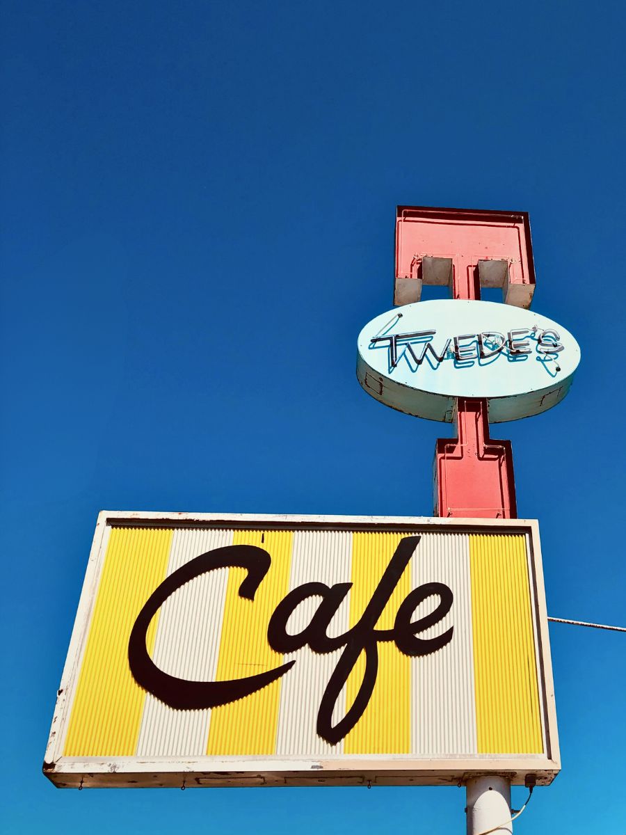 Bright yellow and white vintage cafe sign with the word "Twedes" above it, set against a clear blue sky.