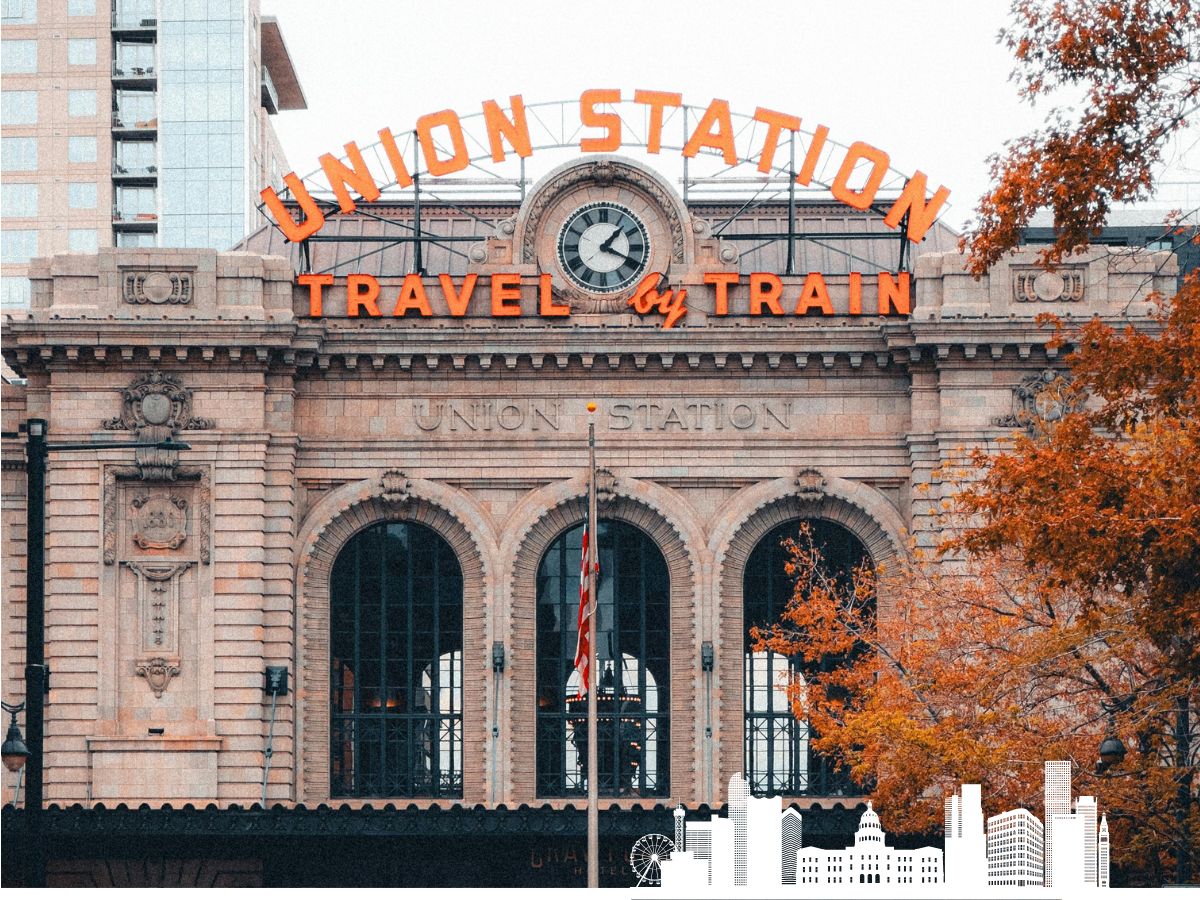 Front view of Union Station with a clock and "Travel by Train" sign, surrounded by urban buildings and autumn trees.