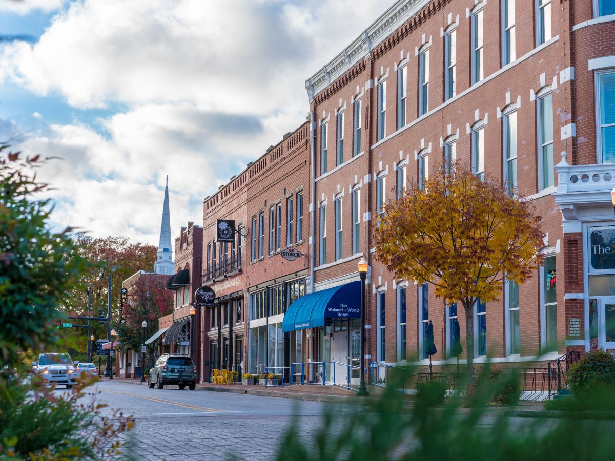 A street view of a small town with red-brick buildings, shops, and autumn trees lining the sidewalk. Cars are parked along the street, and a church steeple is visible in the background.