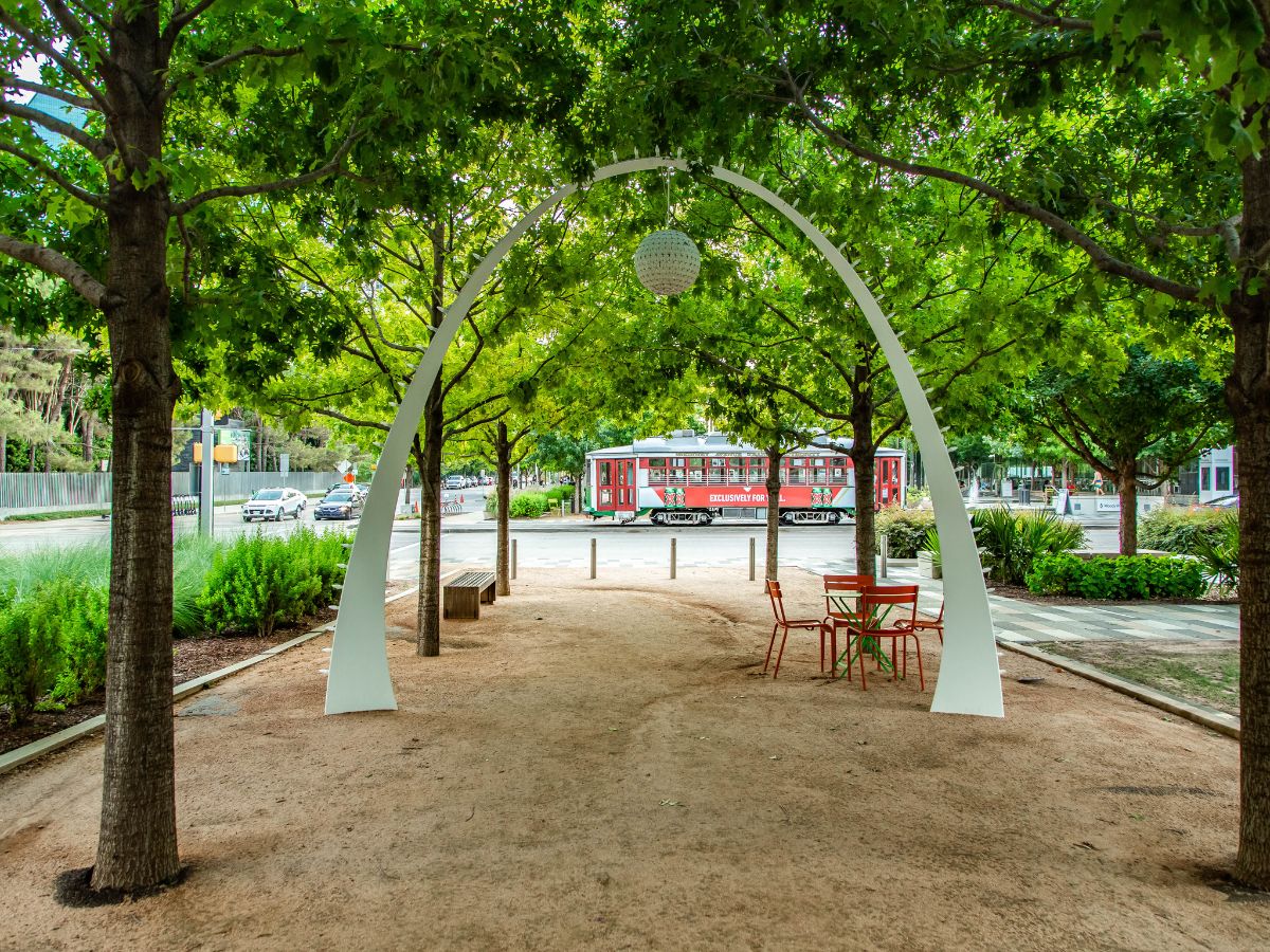 A red streetcar passes by a park with a curved metal archway, trees, benches, and an empty table with chairs.