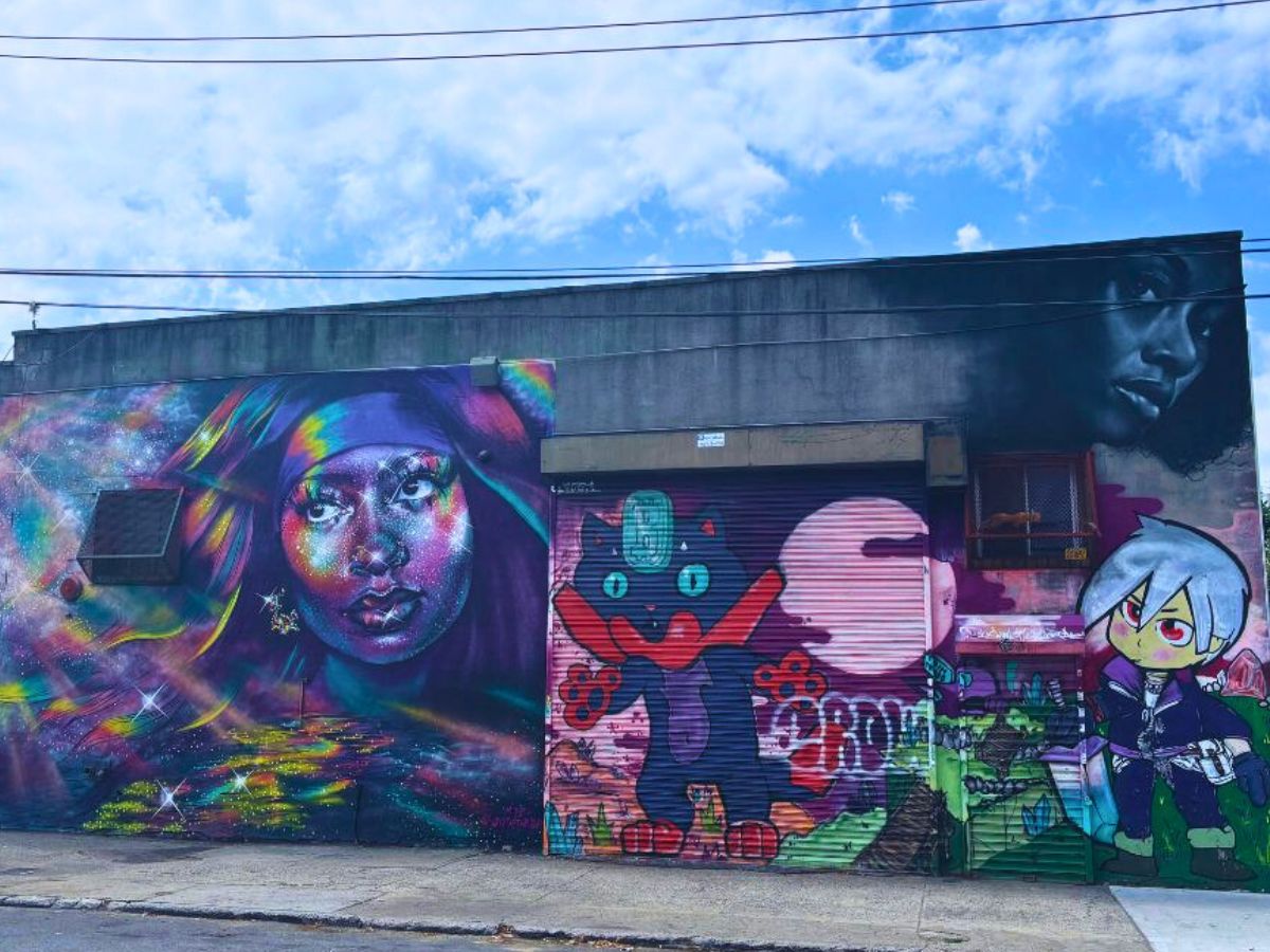 A colorful mural featuring various art styles and characters, painted on the side of a building, with a cloudy sky above.