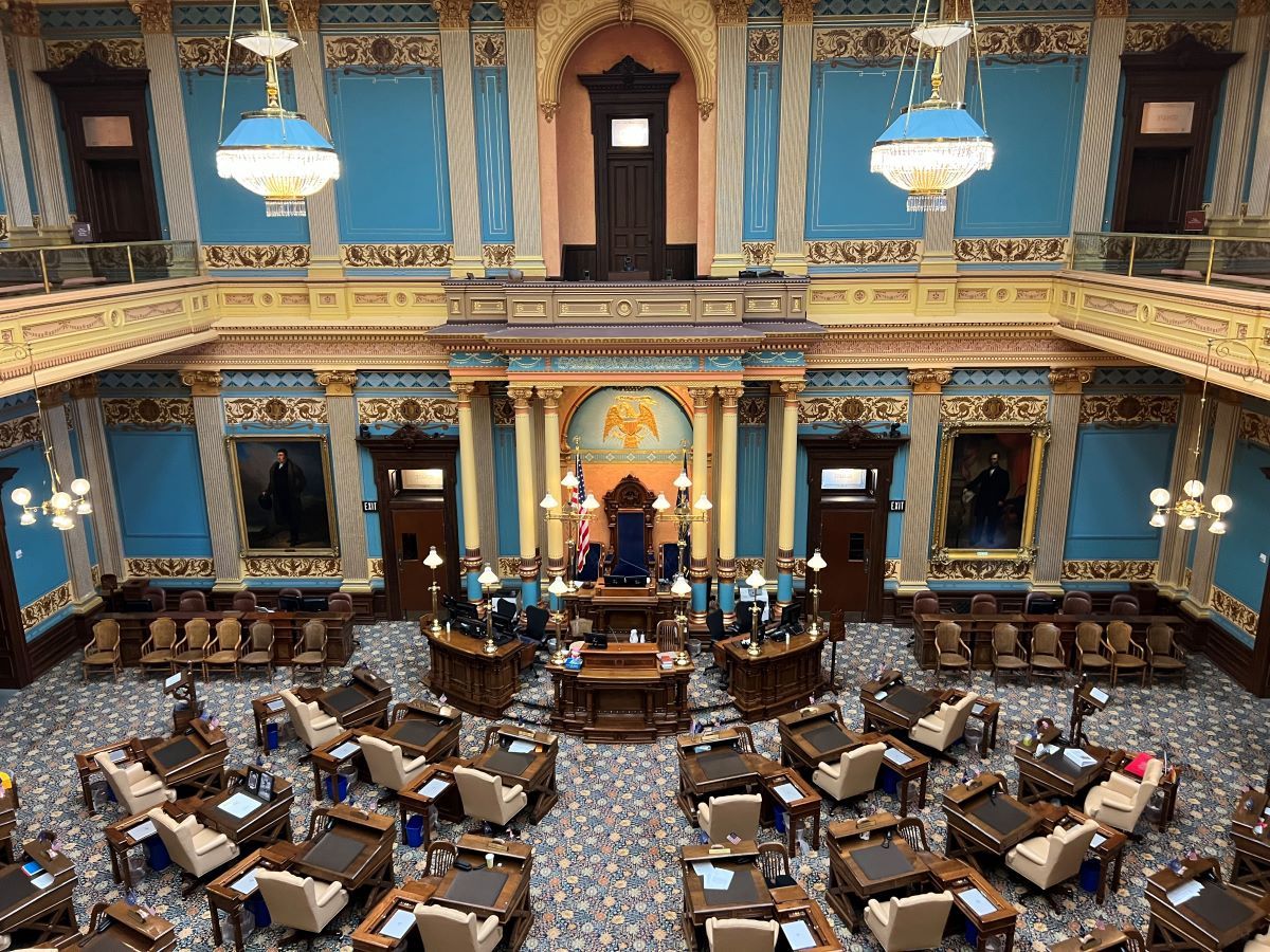 A photograph of a legislative chamber with ornate interior featuring a high ceiling, chandeliers, and a central podium. Desks and chairs for legislators are arranged in the room.
