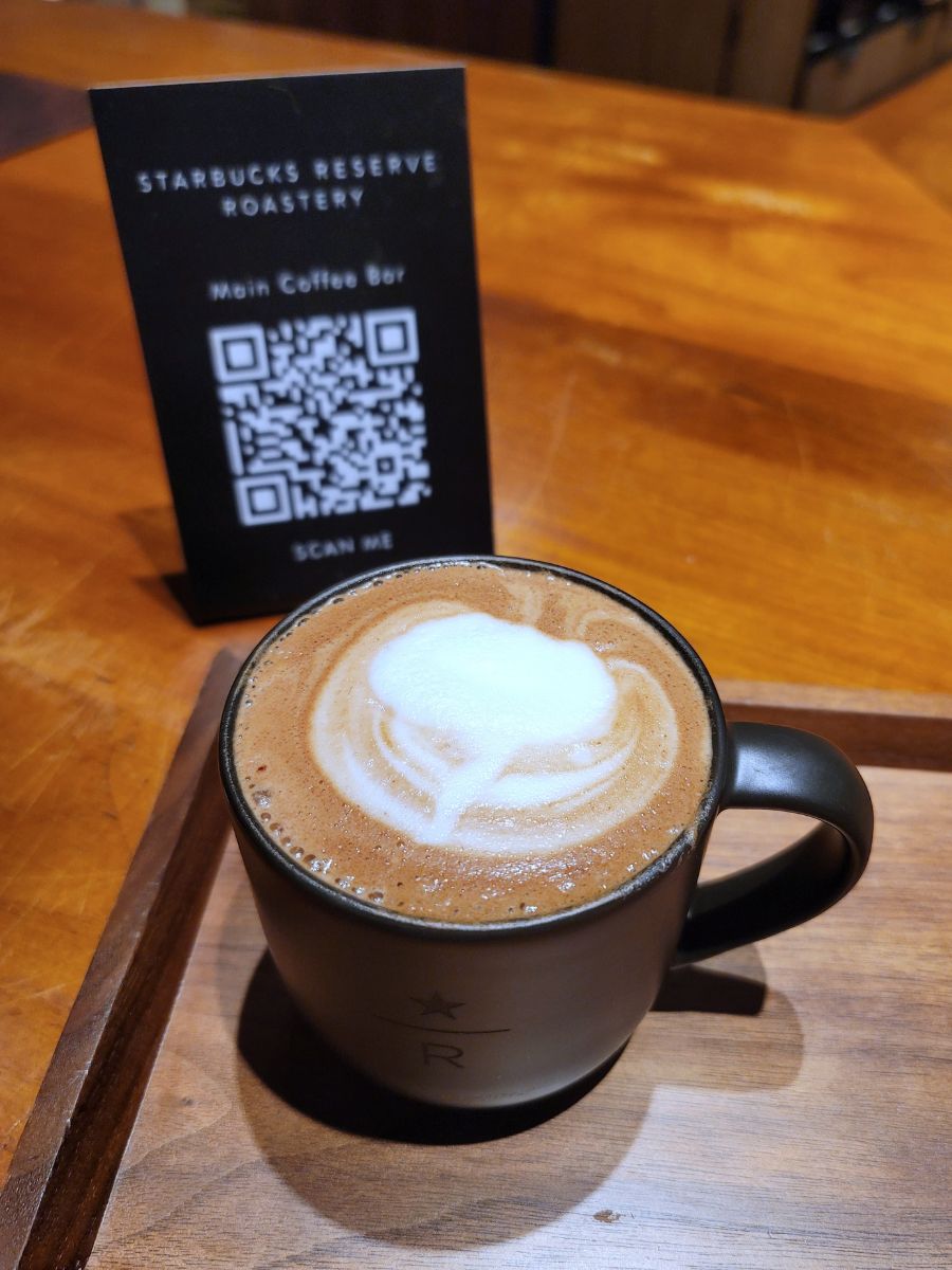 A cup of coffee with latte art sits on a wooden table. In the background, a small sign with a QR code is placed upright, next to a menu that reads "Starbucks Reserve Roastery.