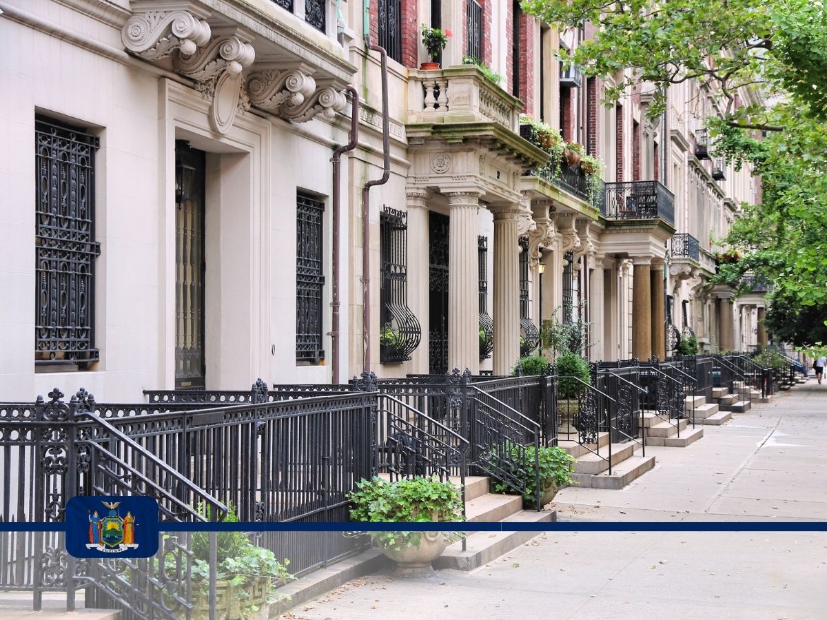 A row of historic, ornate brownstone townhouses with black wrought iron railings and steps in New York. The New York state seal is superimposed on the foreground.