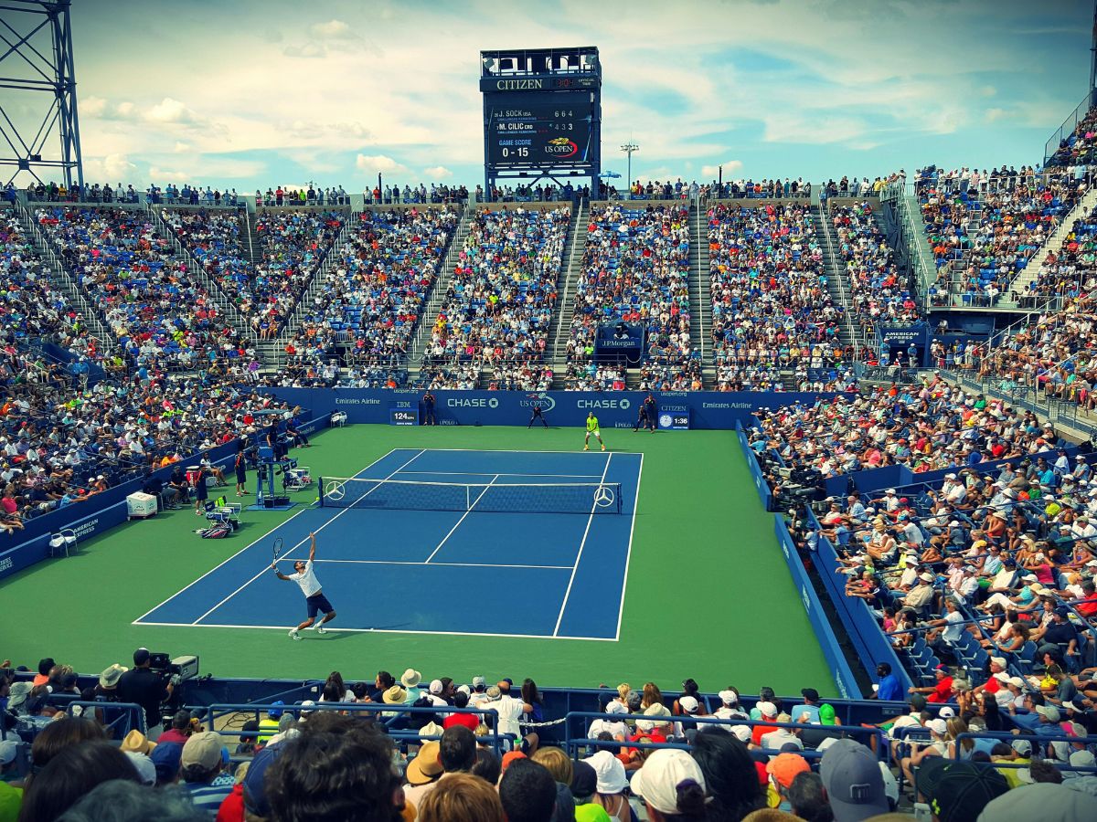 A large crowd watches a live outdoor tennis match on a hard court. One player prepares to serve while the other stands ready. The scoreboard shows the match status.