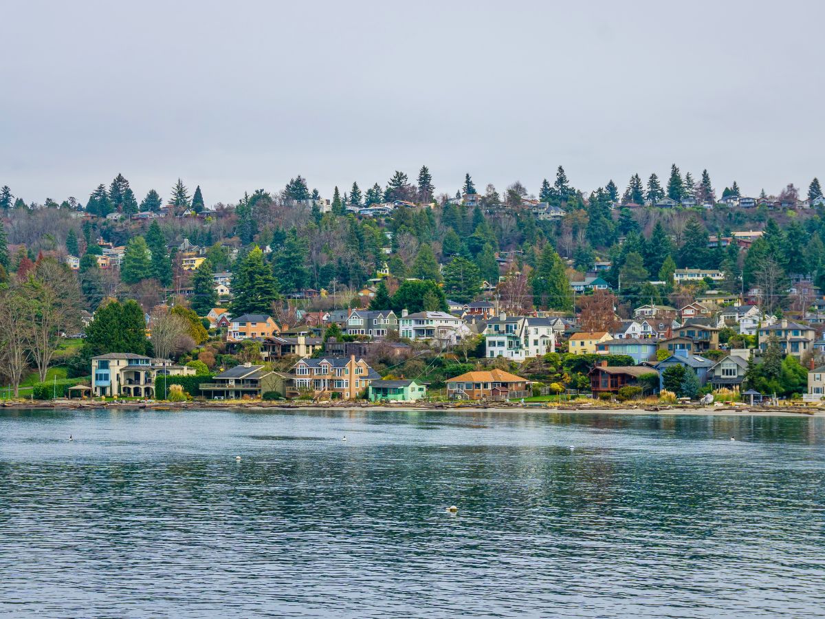 A tranquil waterfront scene with numerous houses nestled among trees on a hillside, reflecting in the calm water below. Overcast sky in the background.