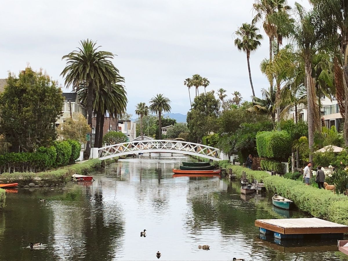 A serene canal lined with greenery, palm trees, and small boats, with a white footbridge spanning the water and cloudy skies overhead.